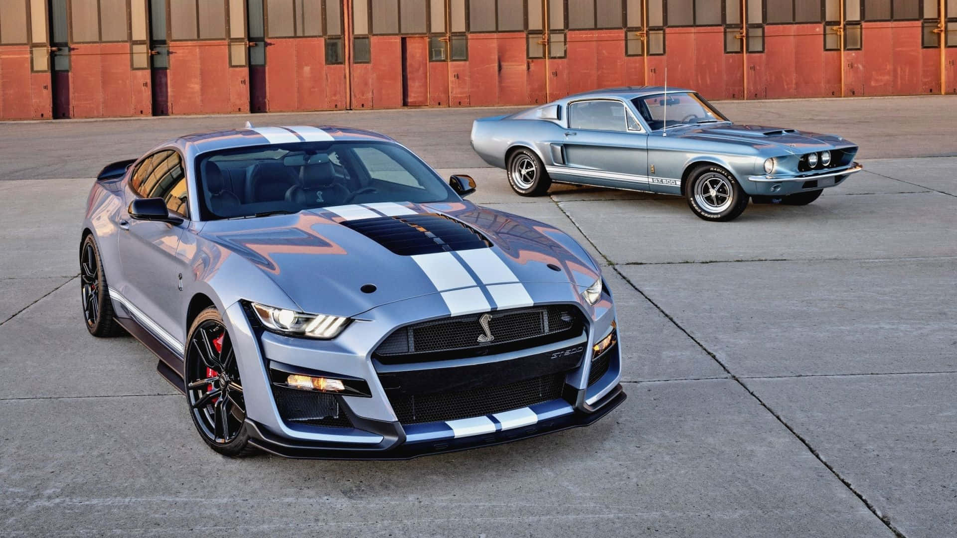  壁紙 — 兩輛帶有白色賽車條紋的銀色Shelby Mustang停泊在紅磚建築前的混凝土上。