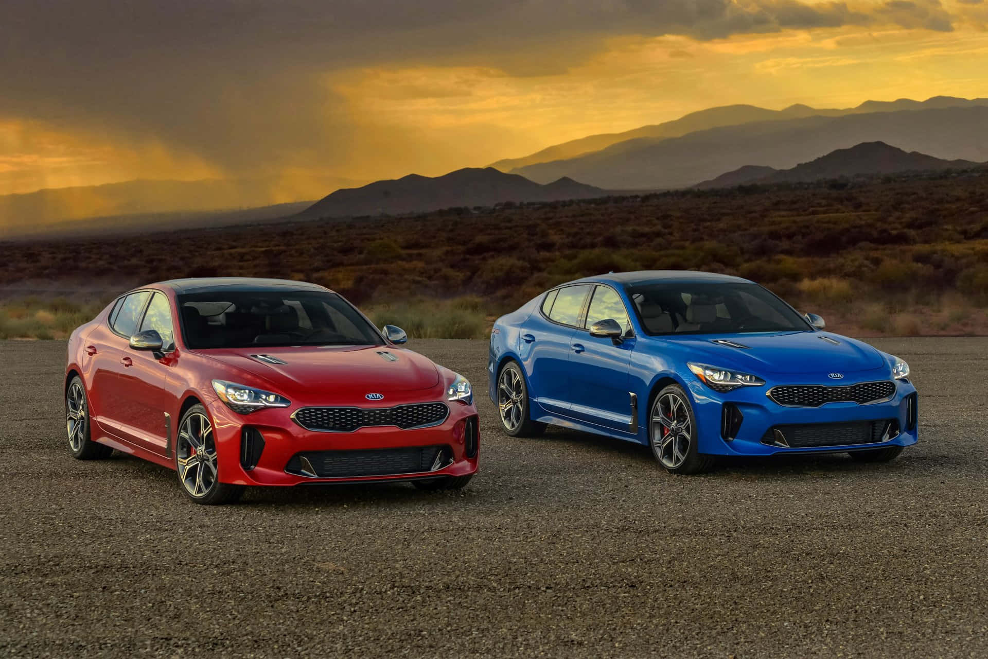 Kia Stinger 桌布 壁紙 — 兩輛時尚跑車紅色和藍色停泊在碎石上，背景是戲劇性的金色日落天空和遠處的山脈。
