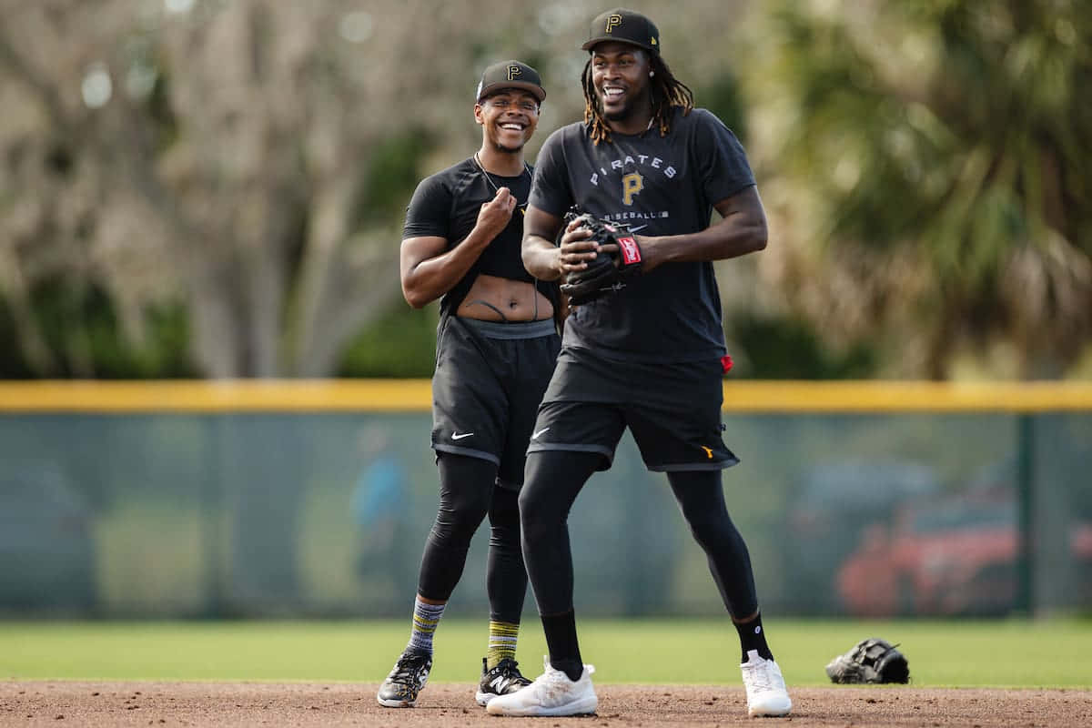  壁紙 — 兩名身穿黑色Pirates裝備的棒球選手面帶笑容，在陽光明媚的場地上一起大笑，背景有樹木。