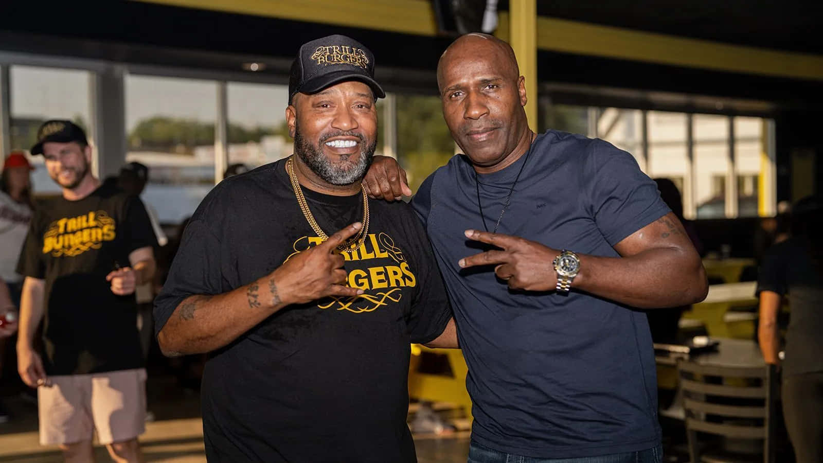  壁紙 — 兩名微笑的男子身穿黑色「Trail Blazers」T恤，在明亮的室內場地以黃色點綴，擺出和平手勢。