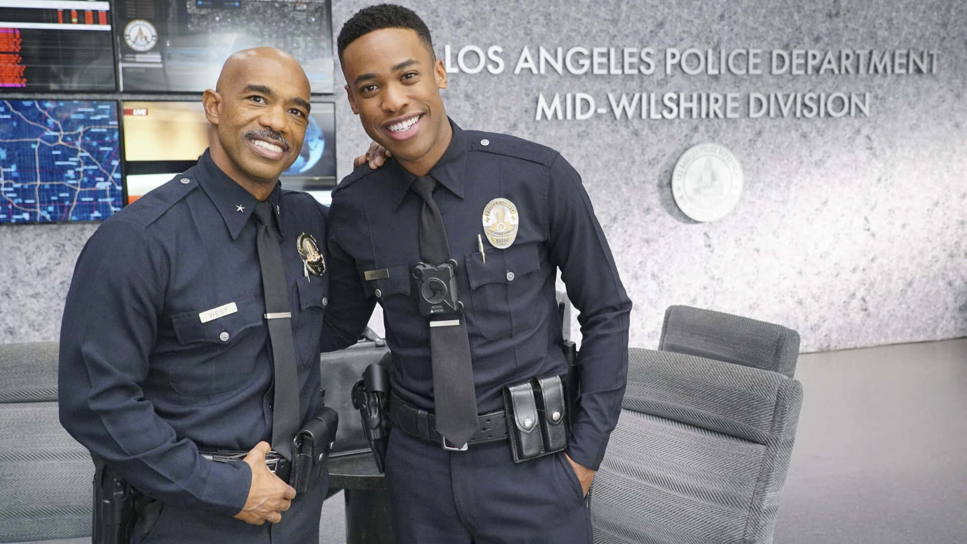 壁紙 — 兩名身穿制服、面帶微笑的警察在現代化的洛杉磯警察局指揮中心自信地擺姿勢，身後有顯示器和部門標誌。