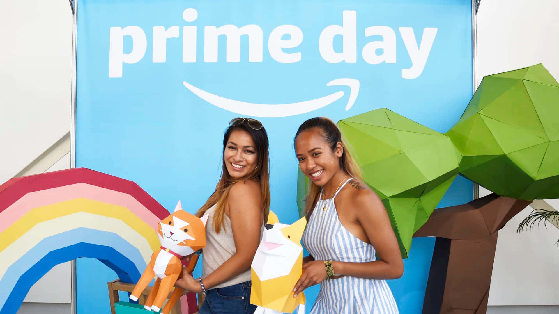  壁紙 — 兩位微笑的女性在明亮的藍色Amazon Prime Day橫幅前，手持色彩繽紛的紙動物擺姿勢。