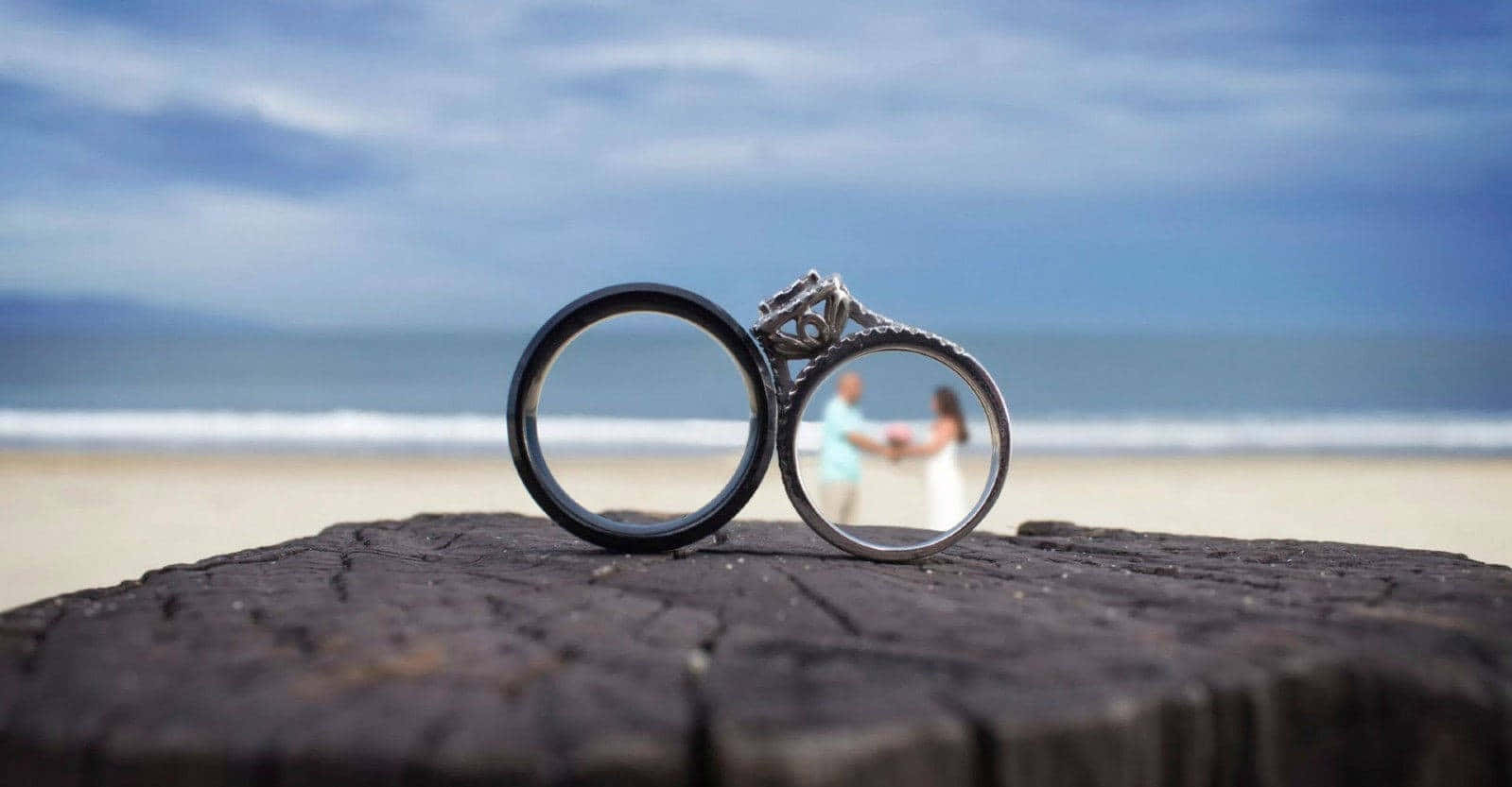 木頭上的兩枚結婚戒指，背後是一對在海灘上牽手的情侶。