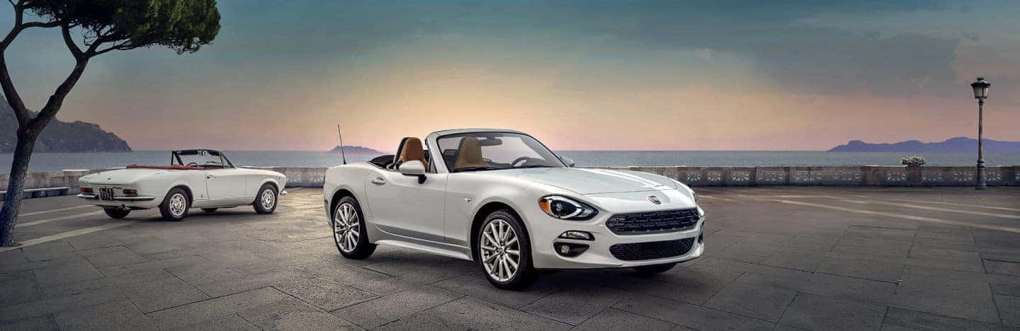 Fiat 124 Spider 桌布 壁紙 — 兩輛白色開篷跑車停泊在石頭步道上，俯瞰日落時平靜的大海。