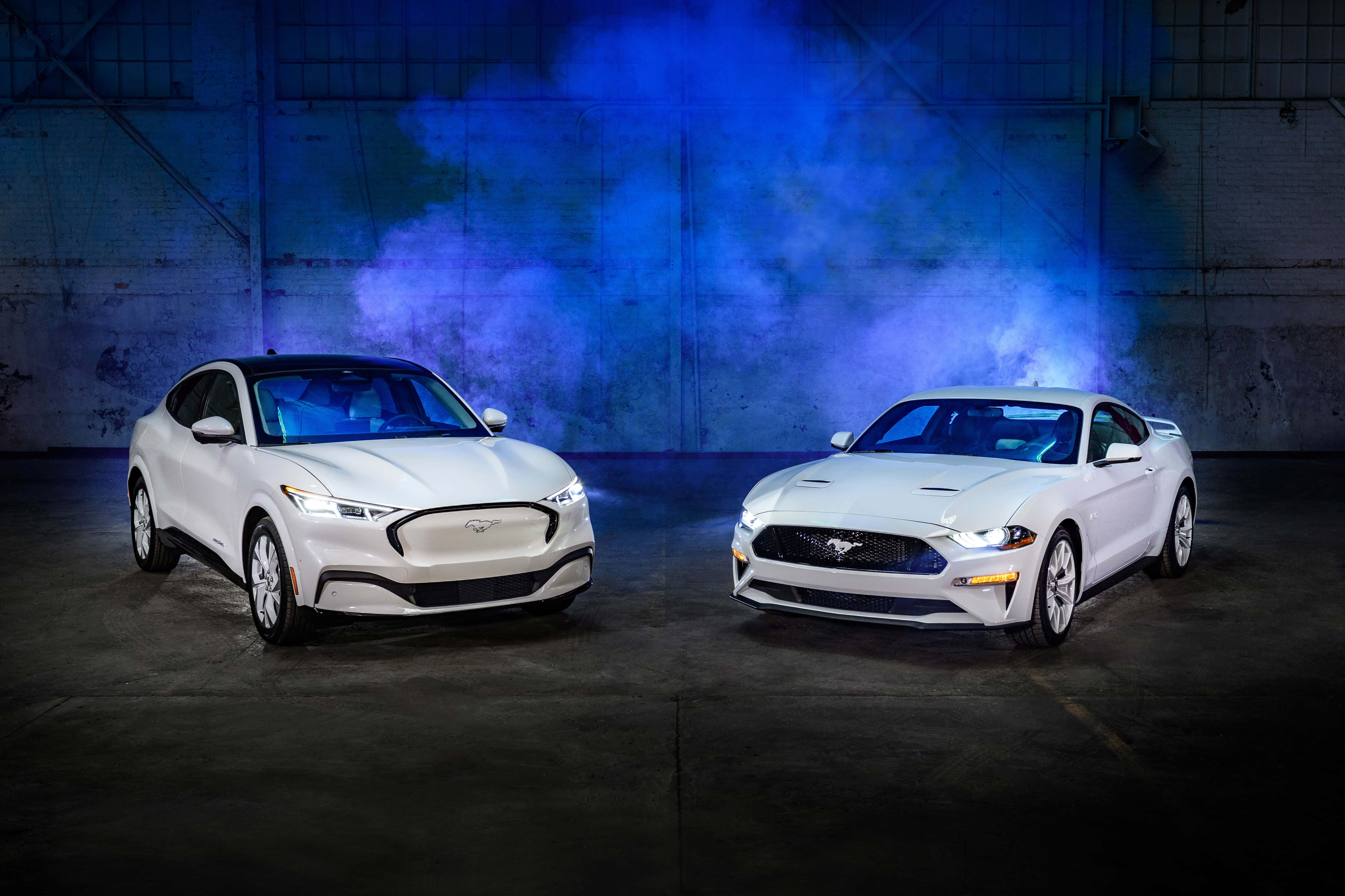 Ford Mustang California Special 桌布 壁紙 — 兩輛白色福特野馬，一輛電動一輛經典，在粗糙的工業車庫中，於戲劇性的藍色煙霧下對峙。