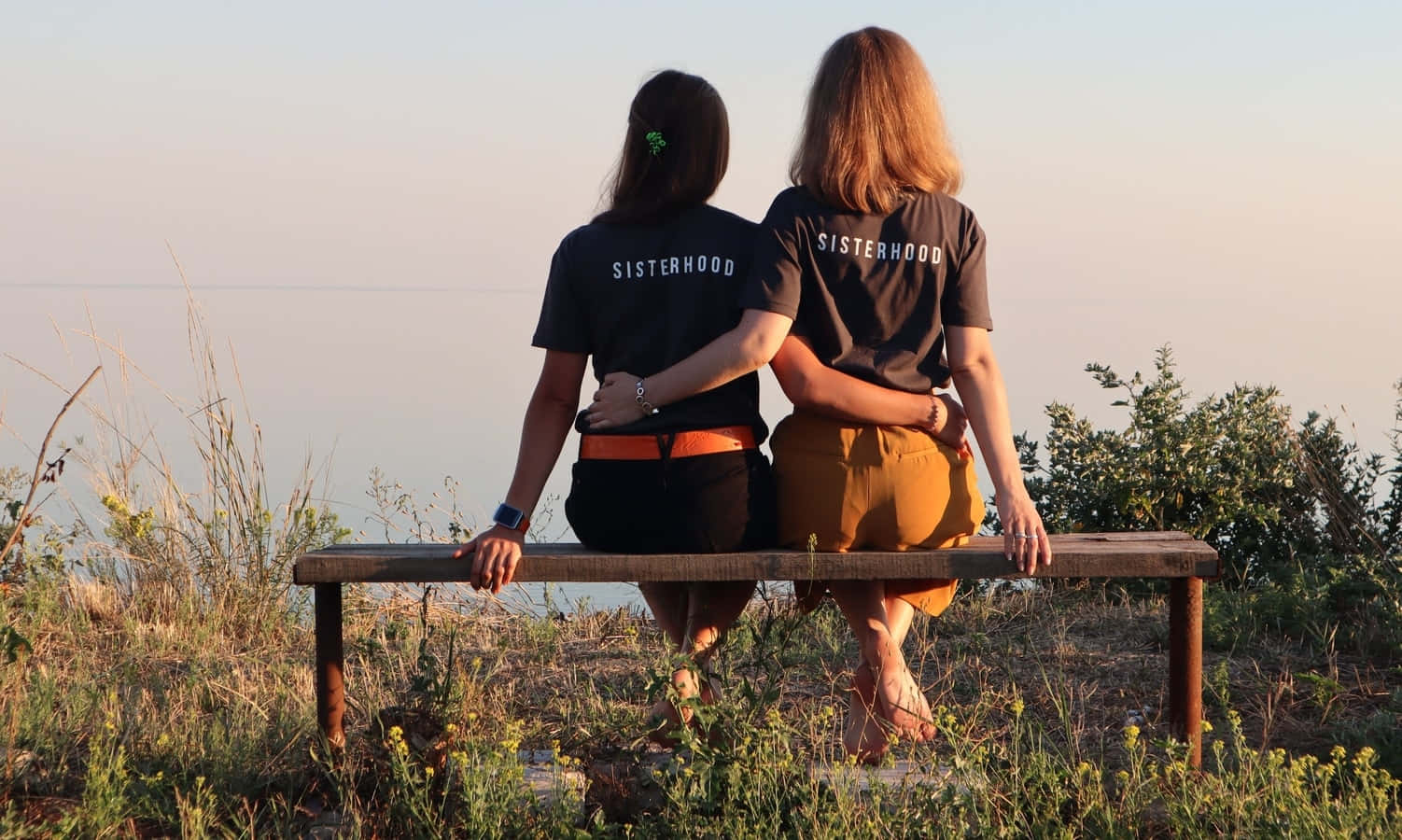  壁紙 — 兩位身穿相配「SISTERHOOD」T恤的女性背對背坐在木長椅上，俯瞰金色時刻的平靜大海。