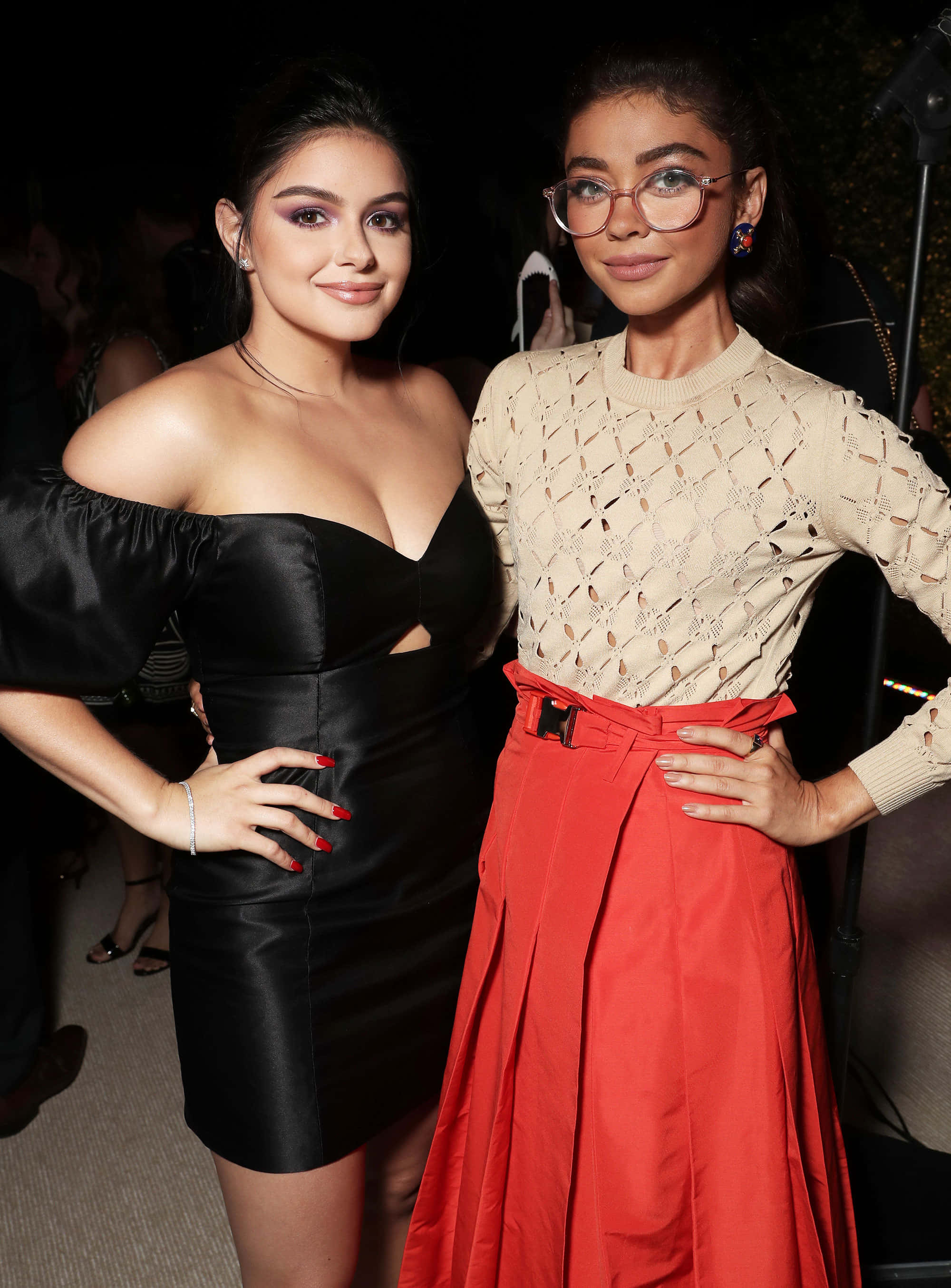 Sarah Hyland 桌布 壁紙 — 兩位女性在華麗活動中自信擺姿勢，一位身穿黑色緞面連衣裙，另一位身穿米色針織上衣和紅色裙子。
