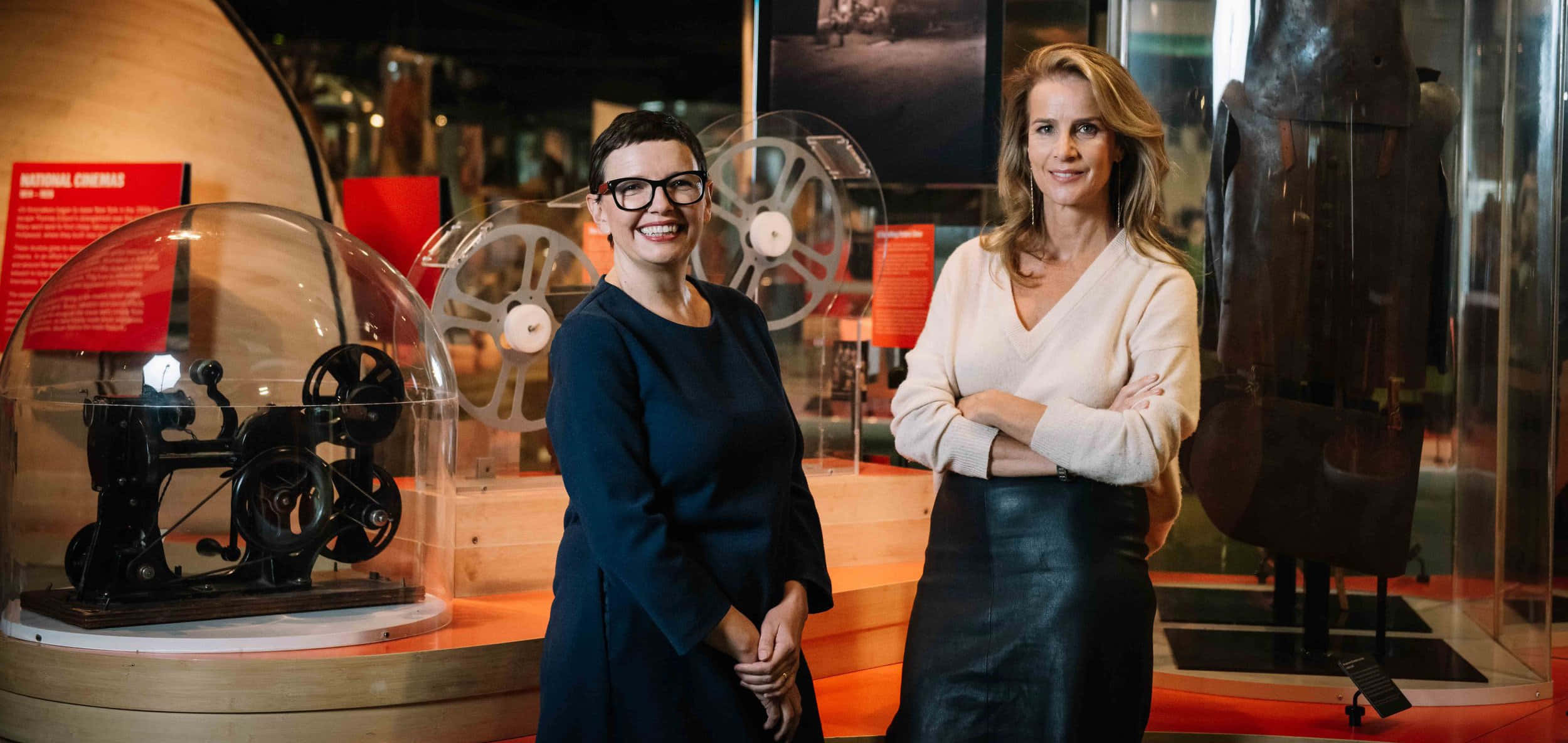 Rachel Griffiths 桌布 壁紙 — 兩位女士在溫暖燈光下的博物館展覽中，自信地微笑站在復古電影設備旁。