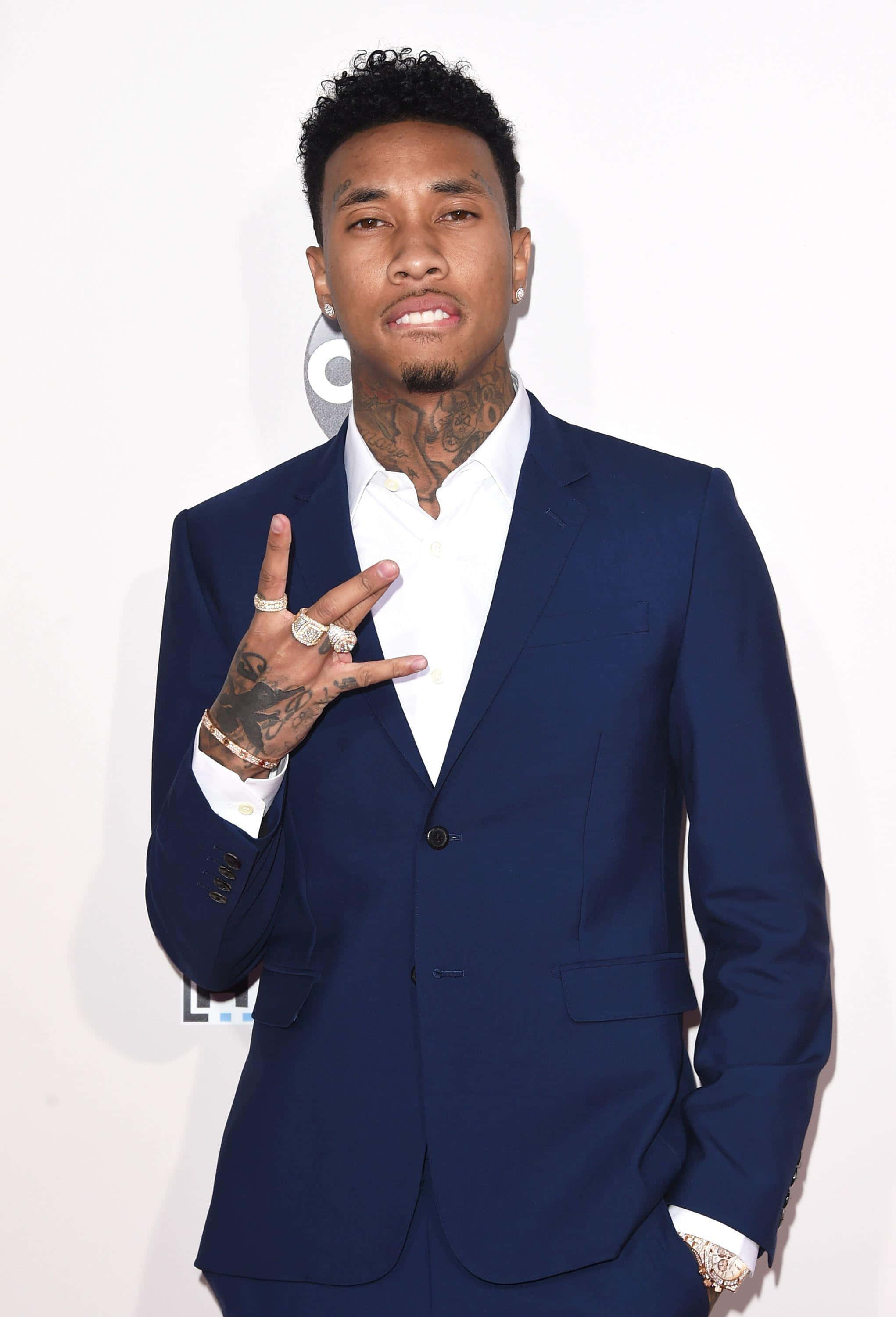 Tyga 現場舞台表演 壁紙 — 一名身穿海軍藍西裝、帶有紋身和戒指的男子在白色背景前自信地擺姿勢。