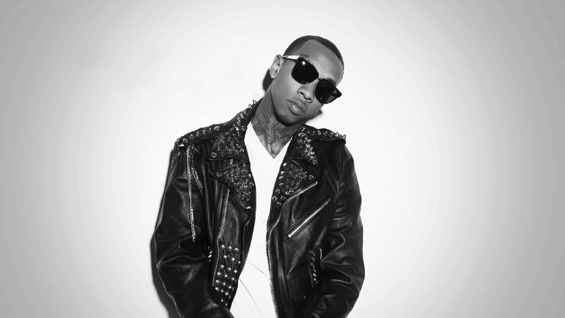 Tyga 舞台表演戴紅帽 壁紙 — 一位時尚男士穿著鉚釘皮褸和太陽眼鏡，在純灰色背景前自信地擺姿勢。