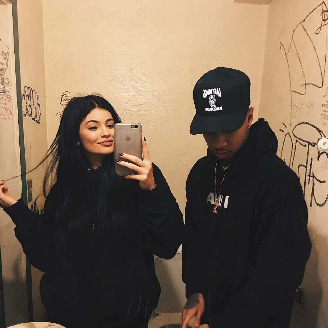 Tyga 與 Kylie 的戀愛模範照片 壁紙 — 兩個穿著黑色連帽衫的人在一個佈滿塗鴉、燈光溫暖的浴室裡對著鏡子自拍。