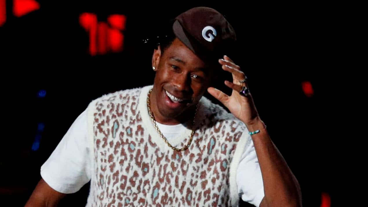Tyler The Creator 大笑頭像 壁紙 — 一個微笑的男人穿著豹紋背心和棕色帽子，在紅色燈光的黑暗背景下調整他的帽子。