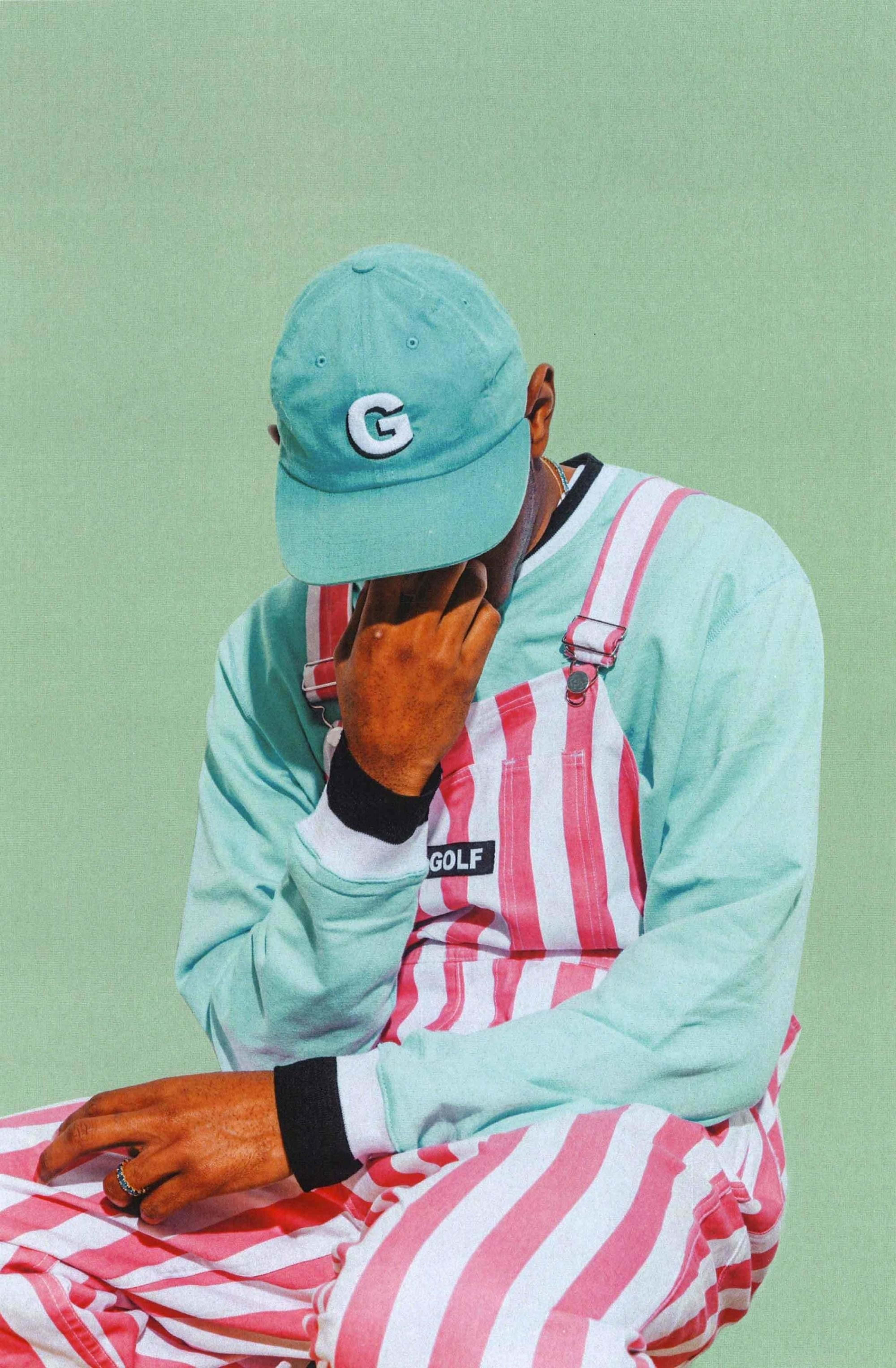 動感視覺 - Tyler The Creator 高解析度影像 壁紙 — 一個人戴著薄荷綠色帽子和粉紅色條紋連身褲，坐在純薄荷綠色背景前，用手遮住臉。