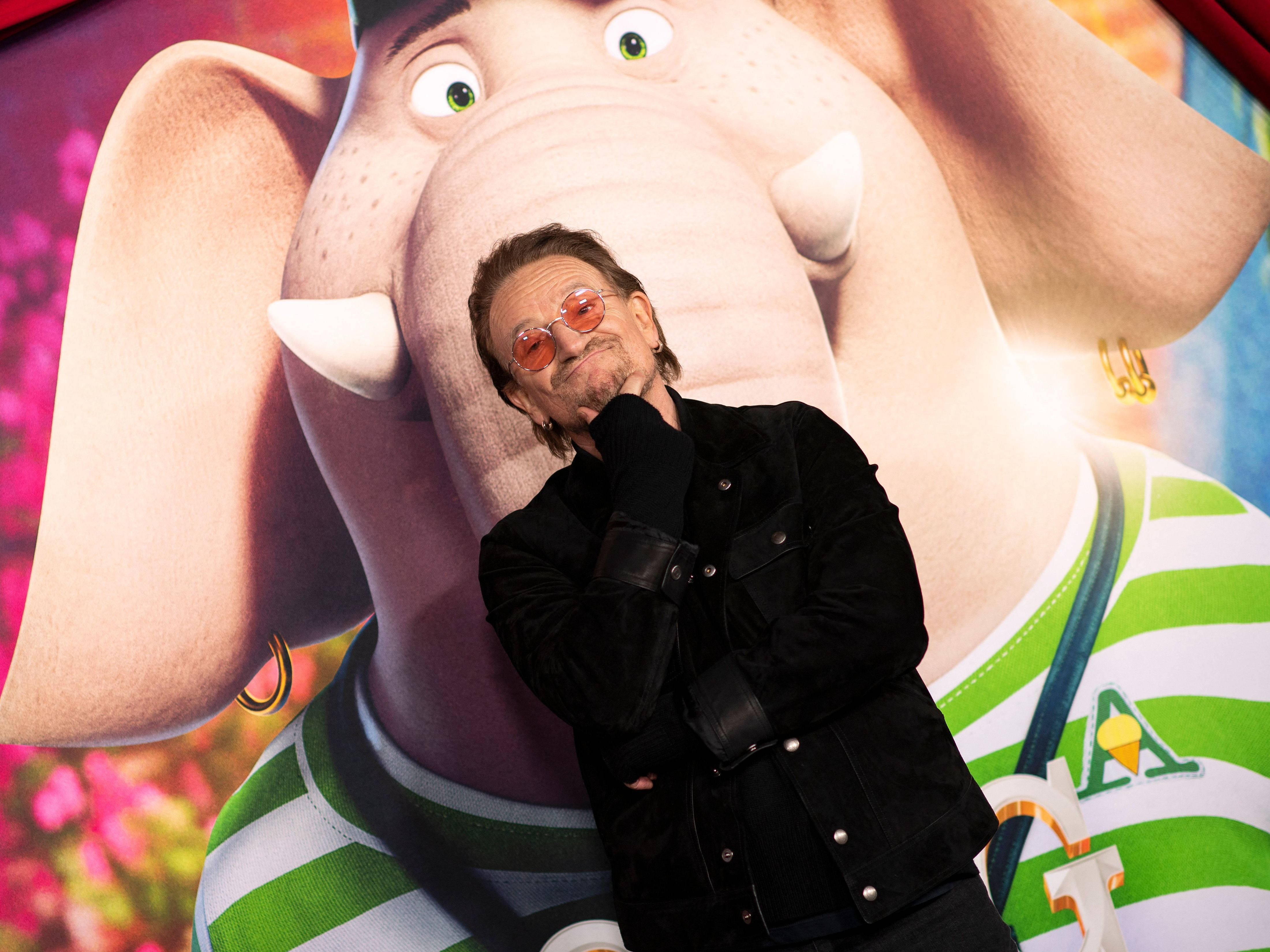 U2主唱Bono為《Sing 2》獻聲配音 壁紙 — 一名戴著太陽眼鏡和黑色外套的男子，自信地在巨大的動畫大象海報前擺姿勢。