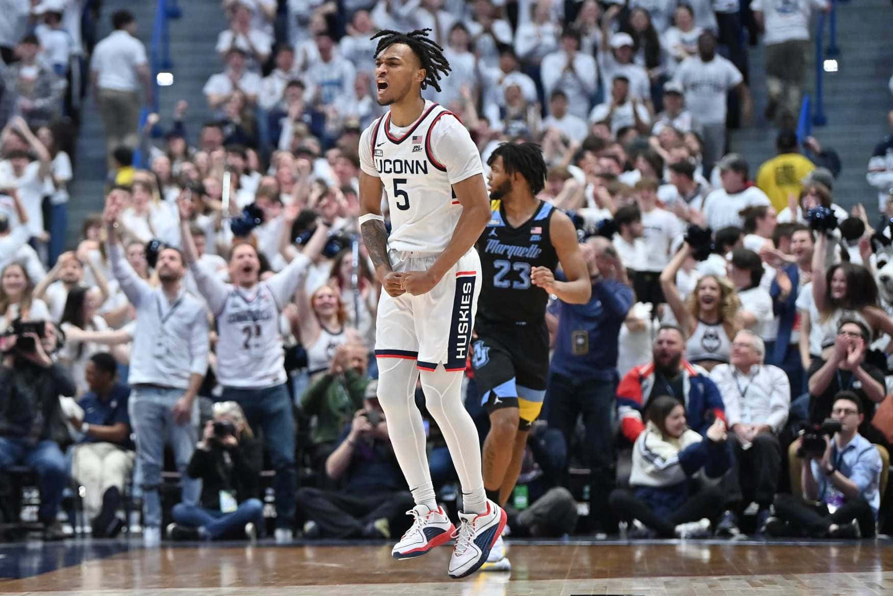  壁紙 — 一位身穿白色球衣5號的UConn籃球運動員在球場上勝利慶祝，身後穿著白色球衣的熱情球迷為他歡呼。