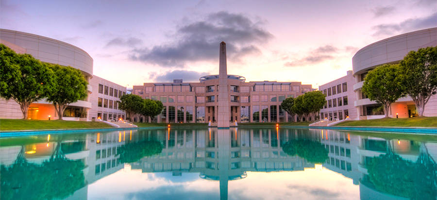 UCSD 校園噴泉風景 壁紙 — 一個現代建築群，擁有曲線建築和中央噴泉，在柔和暮色天空下倒映在平靜的藍綠色水面上。