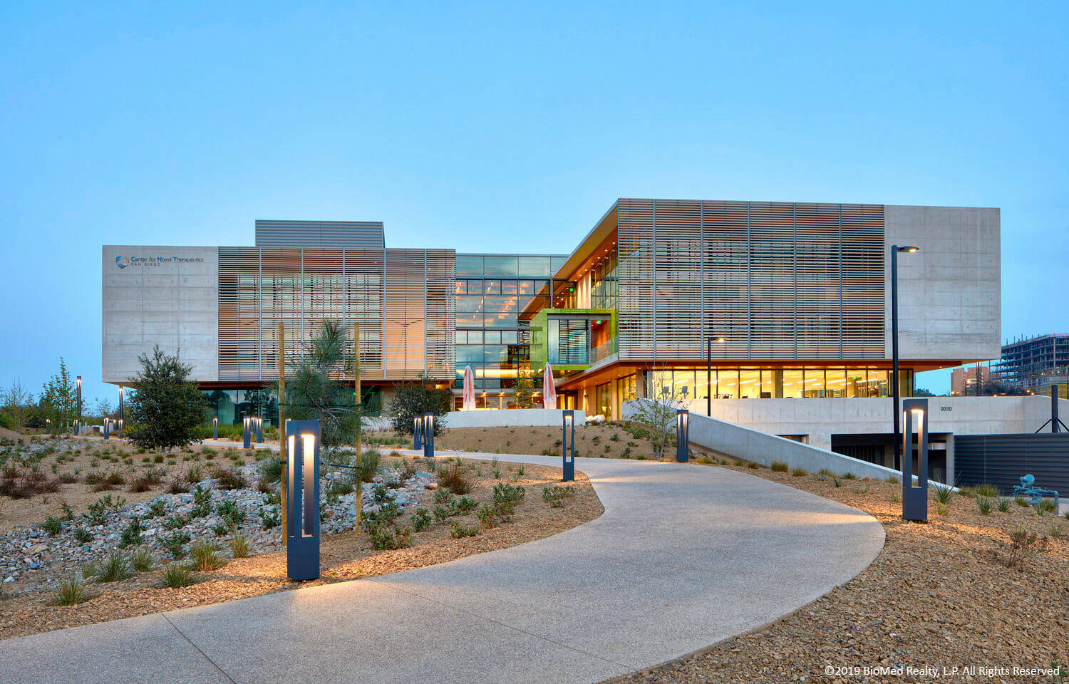 UCSD 沙漠風格建築 壁紙 — 現代建築物，玻璃和木材外牆在黃昏時被照亮，蜿蜒小徑和沙漠景觀。