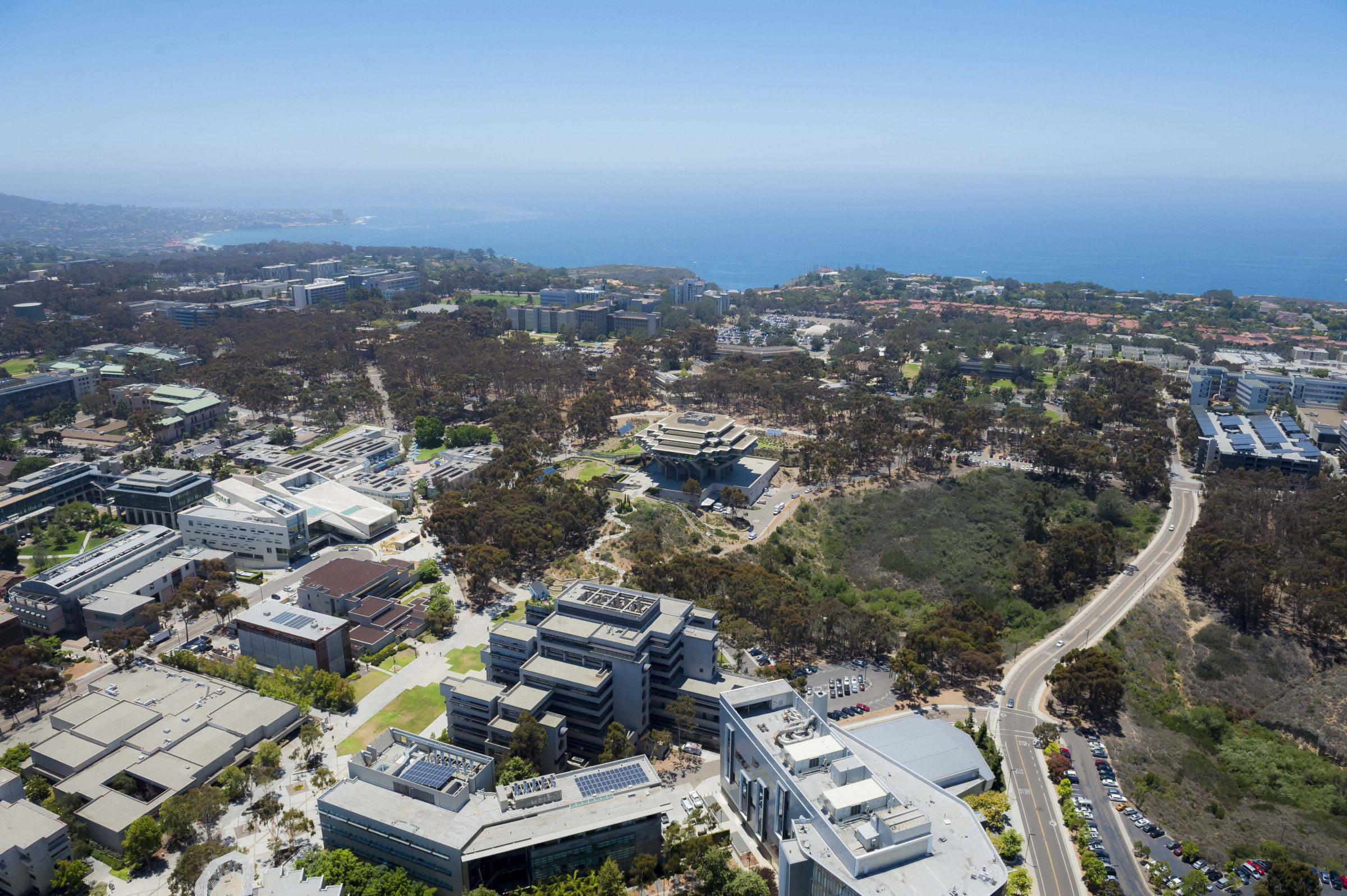UCSD工業校園鳥瞰圖 壁紙 — 現代大學校園的鳥瞰圖，坐落在樹木之間，背景是海洋海岸線，天空湛藍清澈。