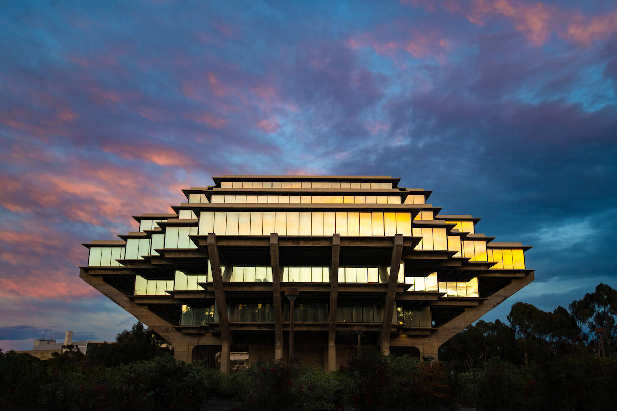 UCSD日落燈光 壁紙 — 一座引人注目的階梯式現代主義建築，在由粉紅色和紫色雲彩繪成的戲劇性暮色天空中溫暖地發光。