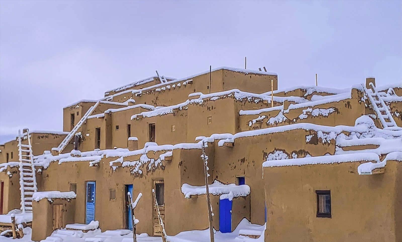 覆蓋白雪的陶斯普ueblo 壁紙 — 白雪覆蓋著藍色門的土坯建築，在蒼白的冬日天空下。