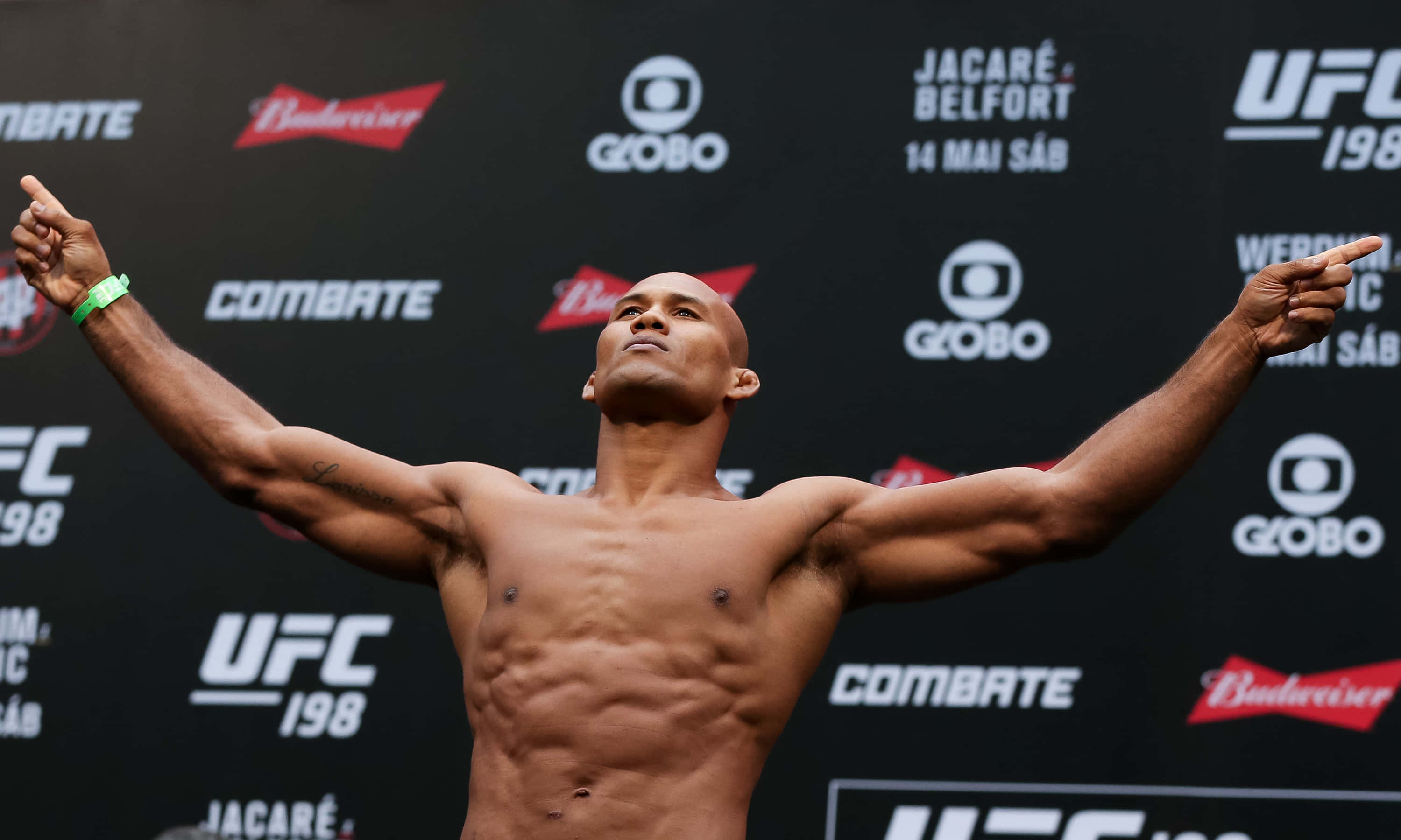 UFC 198賽事稱重 - 羅納爾多·蘇扎 壁紙 — 一名肌肉發達的拳手在UFC過磅活動中赤裸上身，雙臂高舉慶祝勝利，背景為品牌標誌。