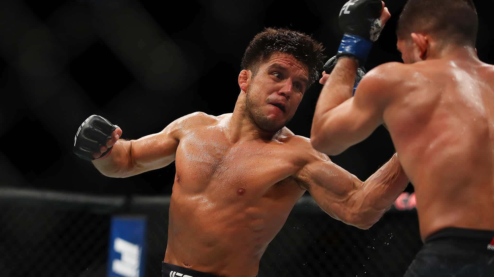亨利·塞胡多於UFC 218小凱撒體育館賽事 壁紙 — 兩名肌肉發達的拳手在鐵籠中激烈交鋒，汗水飛濺，其中一人向對手軀幹擊出強力一拳。