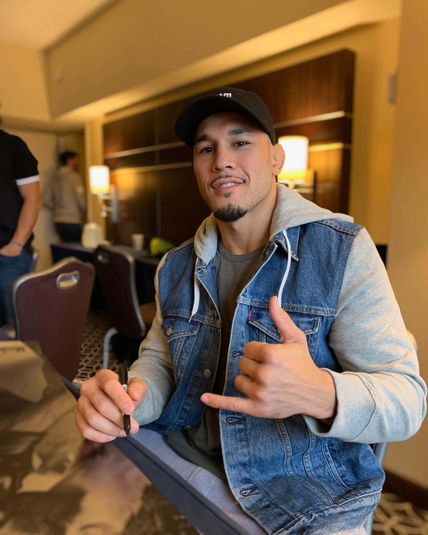 UFC選手布拉德·塔瓦雷斯身穿牛仔外套 壁紙 — 一位微笑的男子穿著牛仔背心和帽子，在燈光溫暖的酒店房間裡舉起大拇指，同時拿著一支筆。