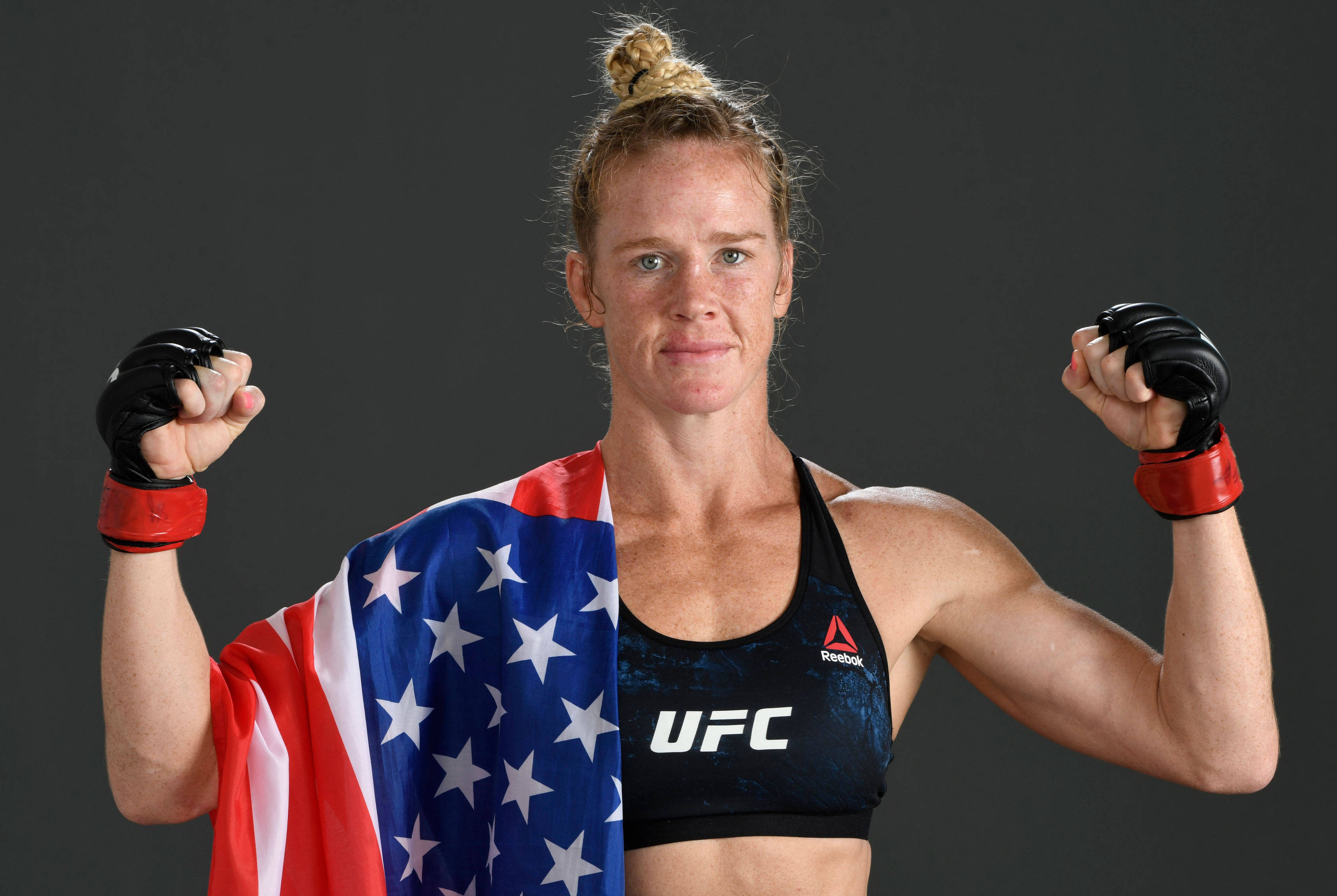 前UFC冠軍 Holly Holm 壁紙 — 一位堅定的女性UFC拳手在黑暗的攝影棚背景下，披著美國國旗展示肌肉。