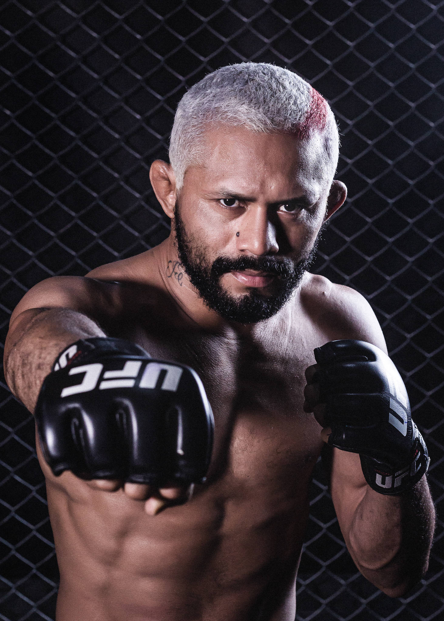 德伊維松·菲格雷多戴UFC手套揮拳 壁紙 — 一位肌肉發達的戰士，銀色頭髮帶有紅色條紋，透過UFC手套在深色鐵絲網前凝視。