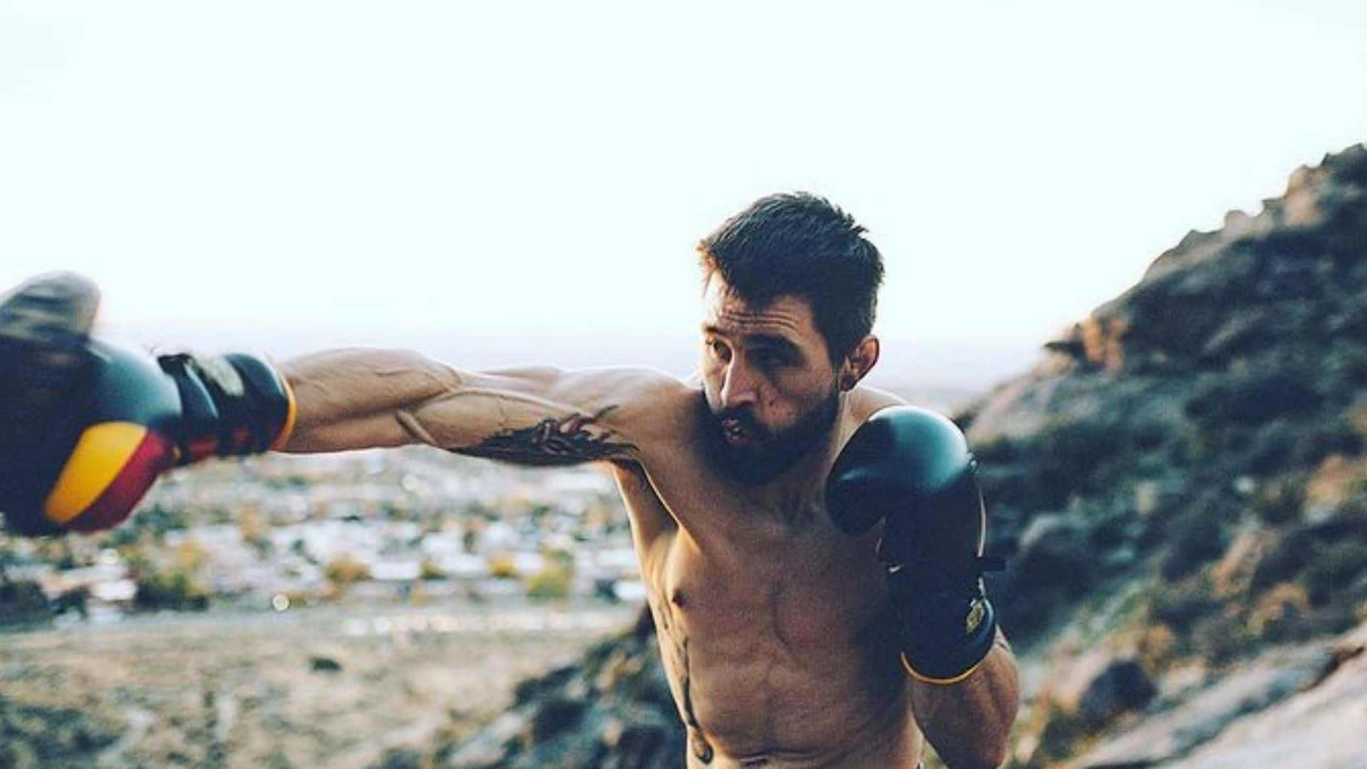 UFC紀錄保持者卡洛斯·康迪特 壁紙 — 一個肌肉發達、留鬍鬚的男人戴住拳擊手套，喺岩石山頂上向住遠方小鎮打出拳，背景係黃金時刻。