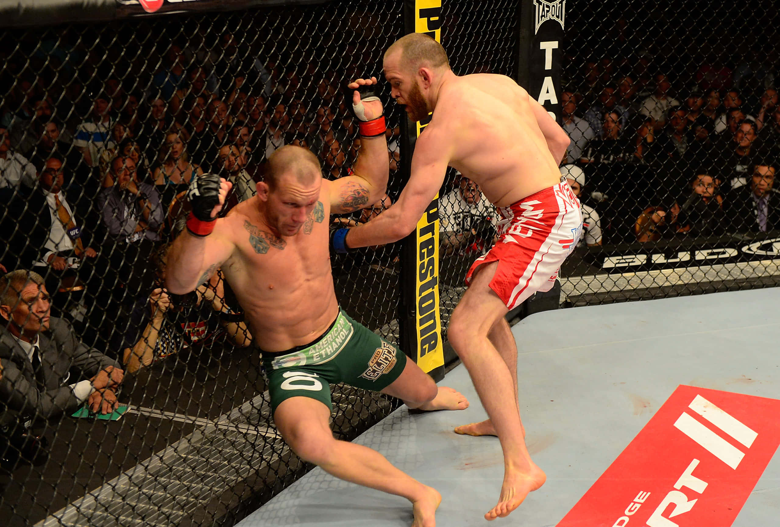 UFC 格雷·梅納德與T.J. 格蘭特對戰 壁紙 — 兩名MMA拳手在八角籠中交鋒，一人使出強力踢擊，另一人在歡呼的人群面前防守。