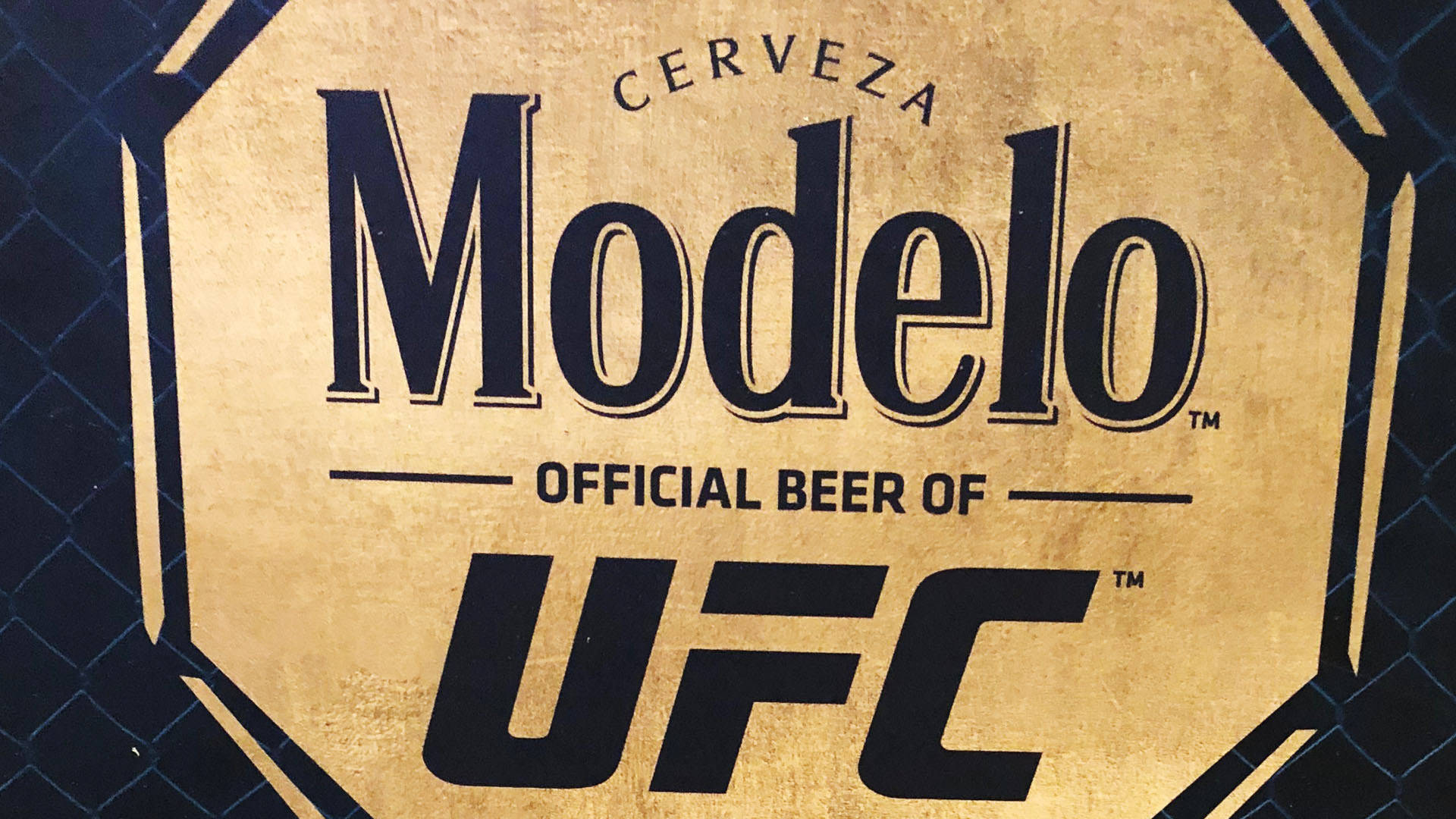 Modelo啤酒標誌，標示為UFC官方啤酒，金色背景帶有鐵絲網圍欄圖案。
