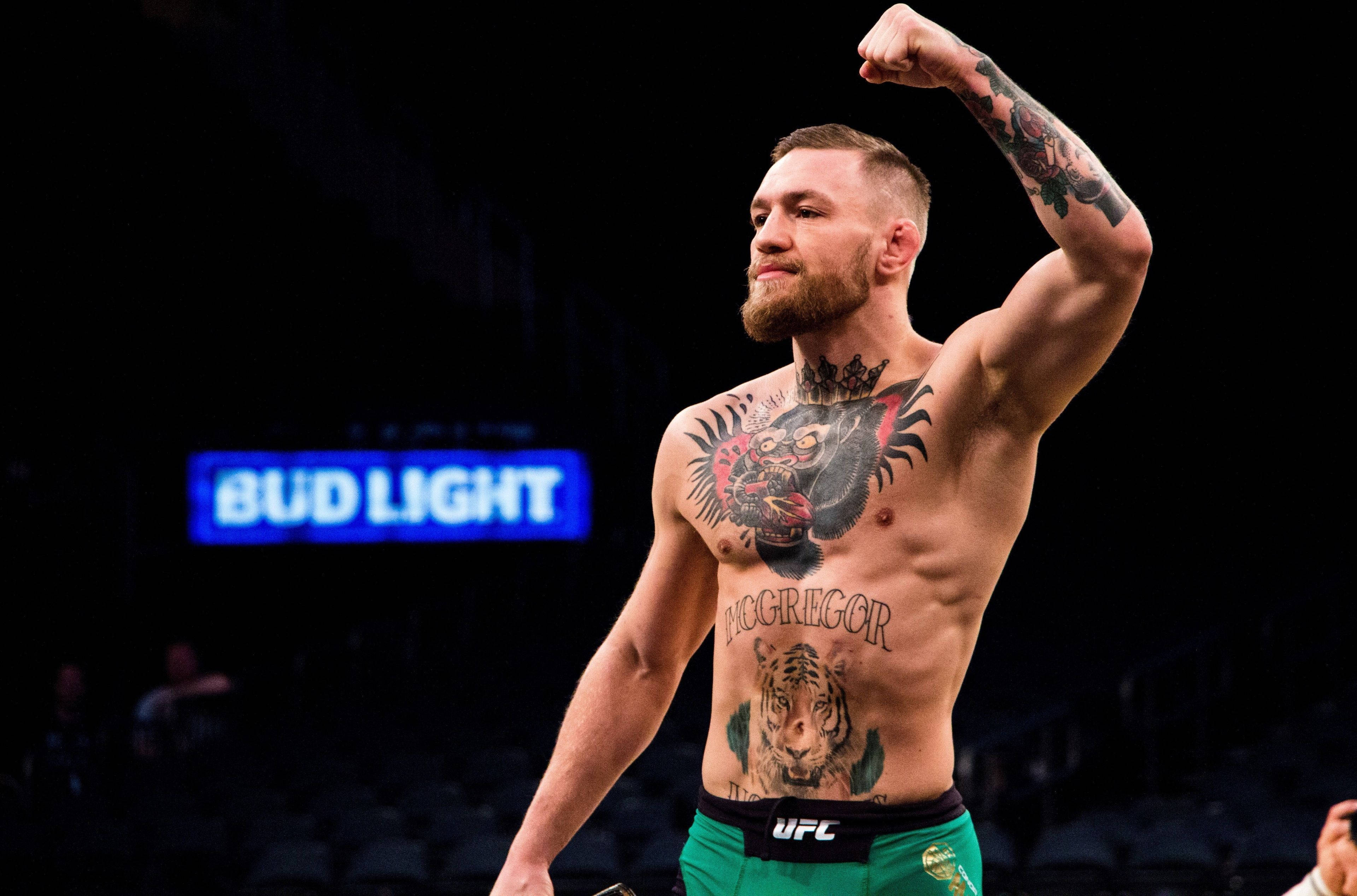 UFC 麥格雷戈舉拳 壁紙 — 康納·麥格雷戈在場館燈光下，身後有Bud Light標誌，以勝利姿態彎曲他帶有紋身的手臂。