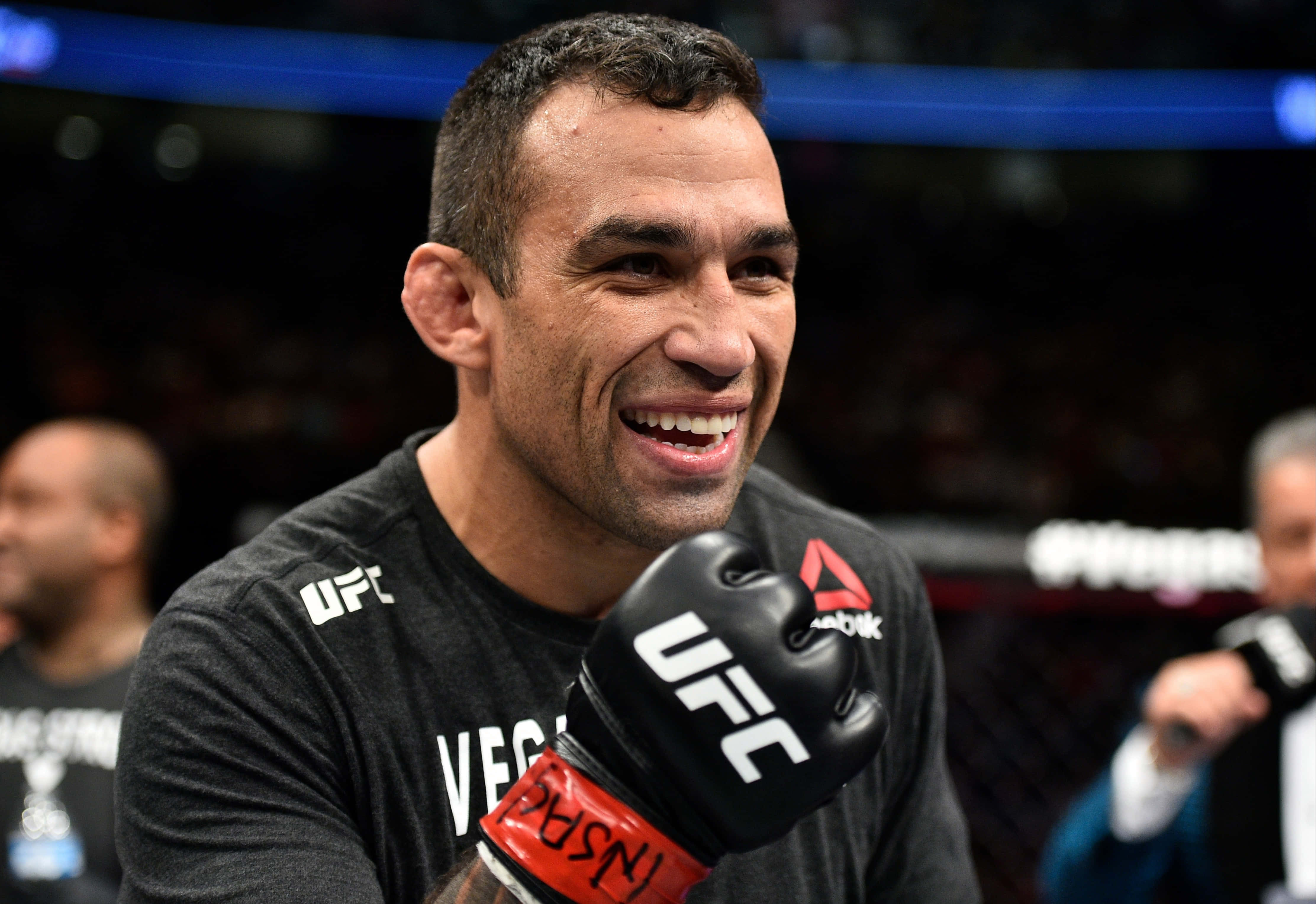 UFC冠軍法比西奧·韋爾杜姆慶祝勝利 壁紙 — 一位微笑的UFC拳手，戴著黑色手套和穿著黑色襯衫，站在八角籠中，身後是模糊的人群和明亮的燈光。