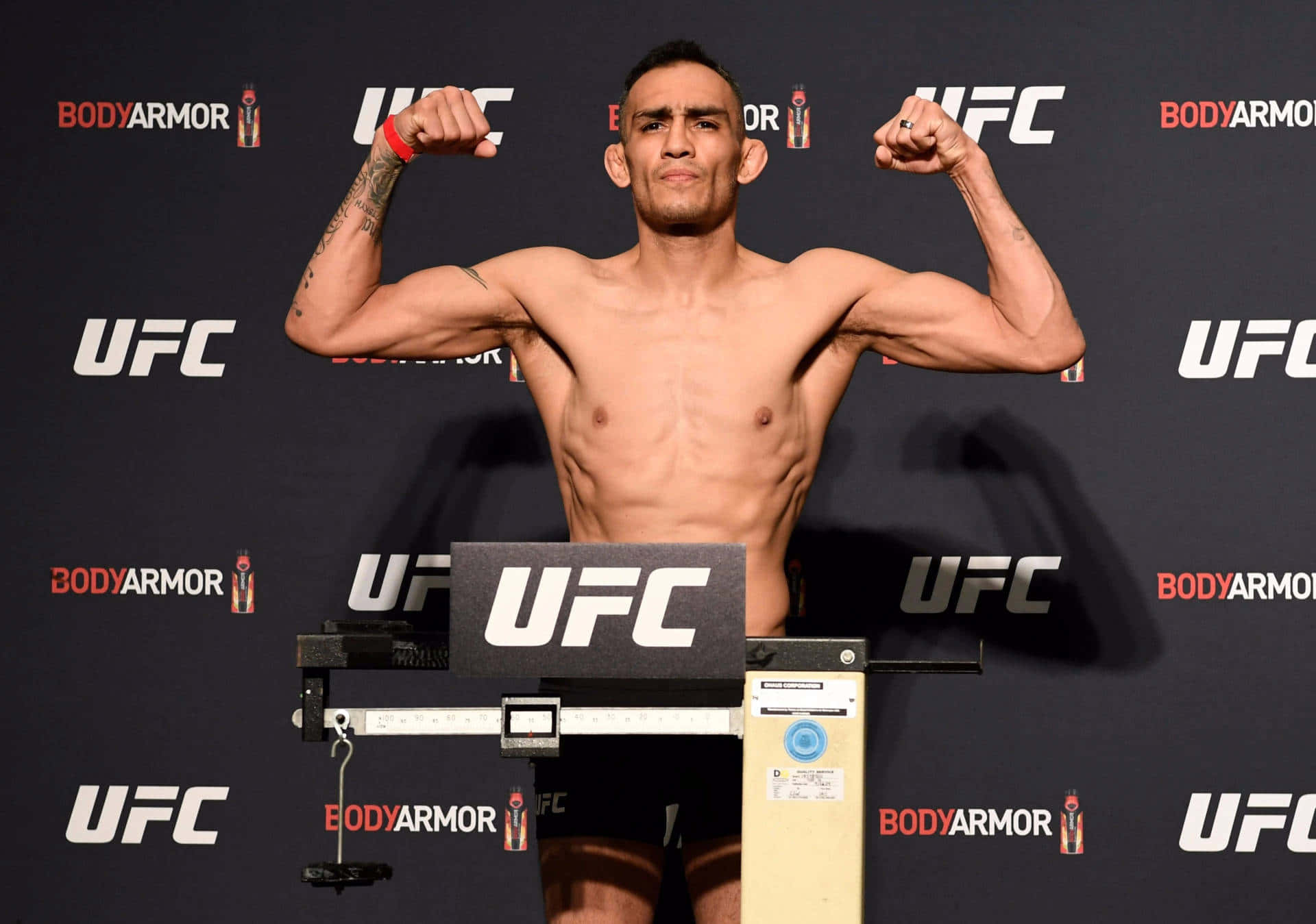 UFC賽事中的自衛技巧示範 壁紙 — 一位肌肉發達的UFC拳手在有品牌背景的稱重活動中，在磅秤上屈起手臂。