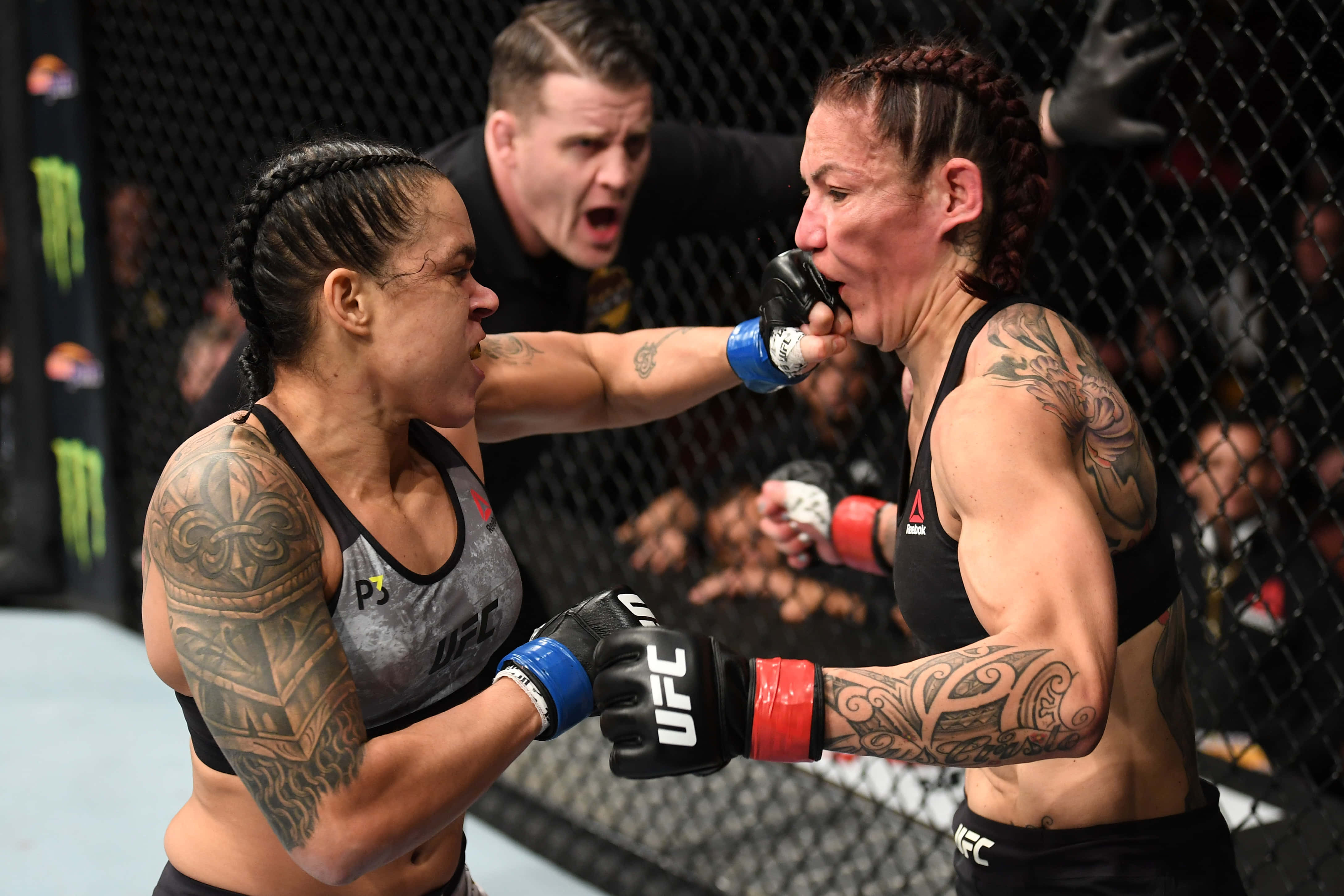 UFC選手Amanda Nunes與Cris Cyborg UFC 232比賽瞬間 壁紙 — 兩名女性MMA拳手在籠中交鋒，身上有紋身、戴著手套，背景中裁判在大喊。
