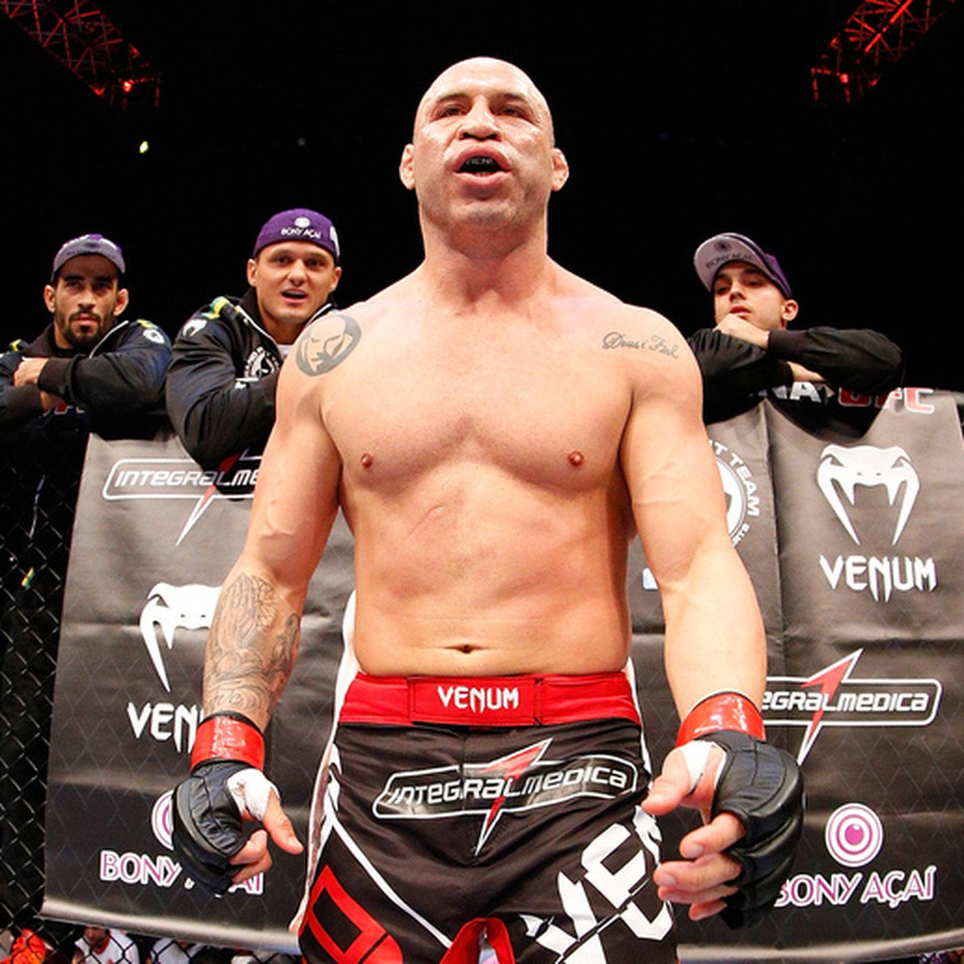 UFC選手Bas Rutten 壁紙 — 一位肌肉發達的MMA拳手穿著紅黑短褲，在八角籠中與隊友一起勝利站立。
