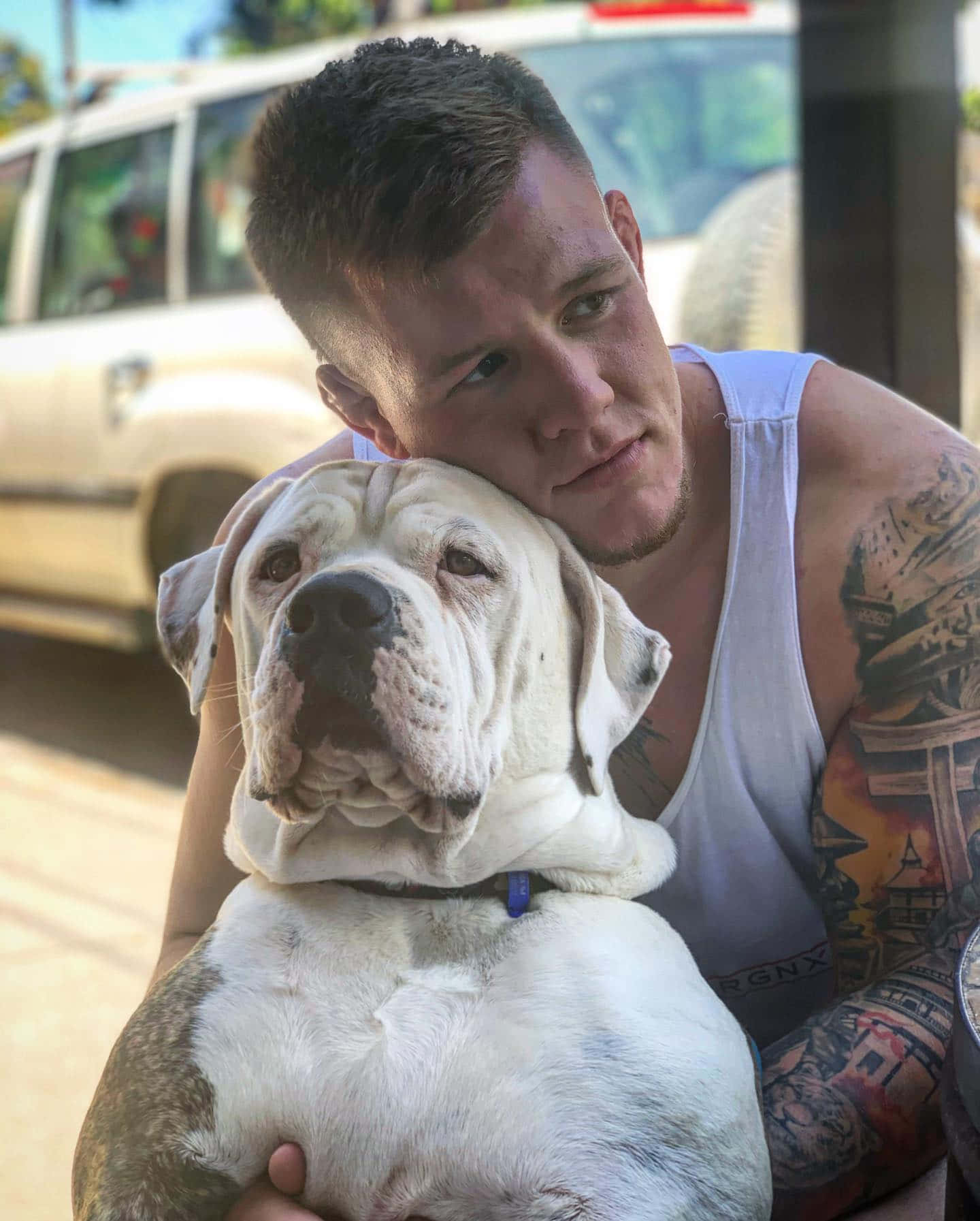 有紋身的男子在陽光明媚的街道上擁抱白色鬥牛犬。