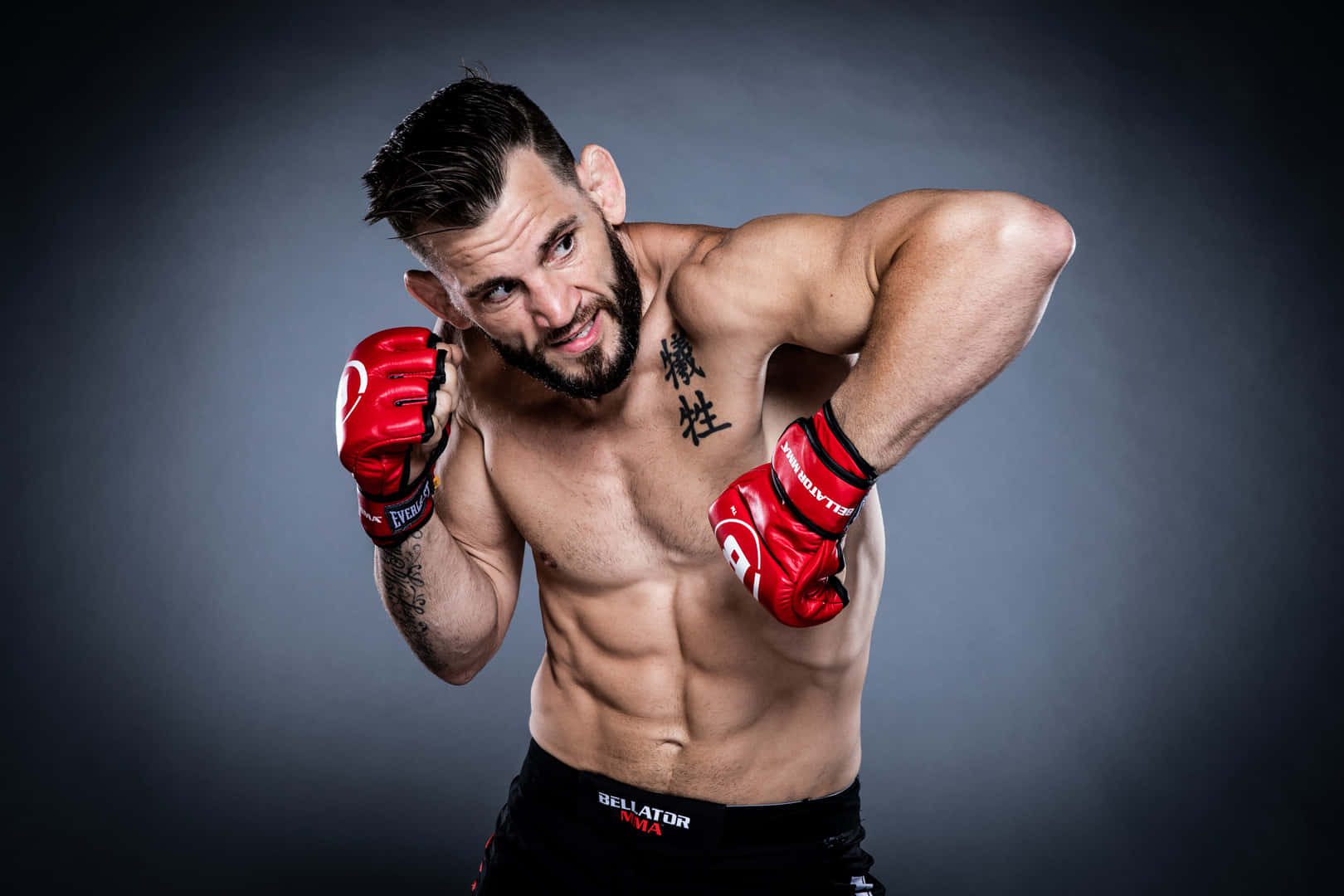 UFC選手Jon Fitch在擂台發起肘擊 壁紙 — 一位留鬍鬚的肌肉發達戰士，戴著紅色手套，在深灰色工作室背景前激烈擺姿勢。