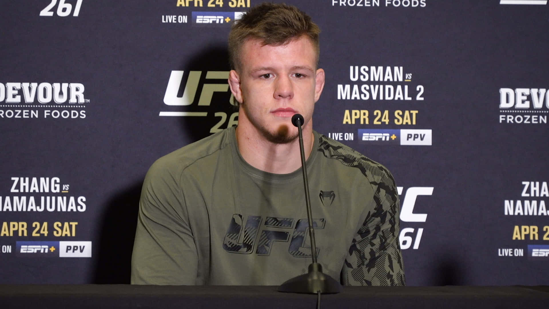 UFC選手吉米·克魯特手持麥克風 壁紙 — 一位專注的UFC拳手坐在記者會的桌子前，面前有麥克風，背景是品牌標誌。