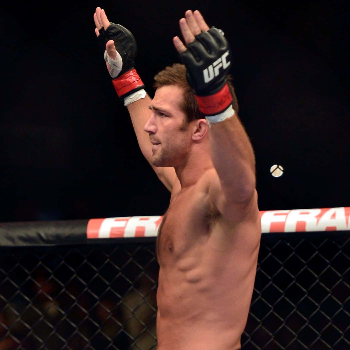 UFC選手盧克·羅克霍德側角鏡頭 壁紙 — 一位肌肉發達的UFC拳手在昏暗的八角籠內舉起戴著手套的雙手慶祝勝利。
