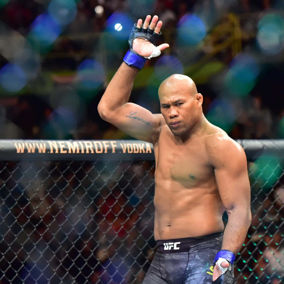 UFC選手羅納爾多·蘇薩高舉右手 壁紙 — 一位肌肉發達的拳手在UFC八角籠內向粉絲揮手，背景是模糊的人群燈光。