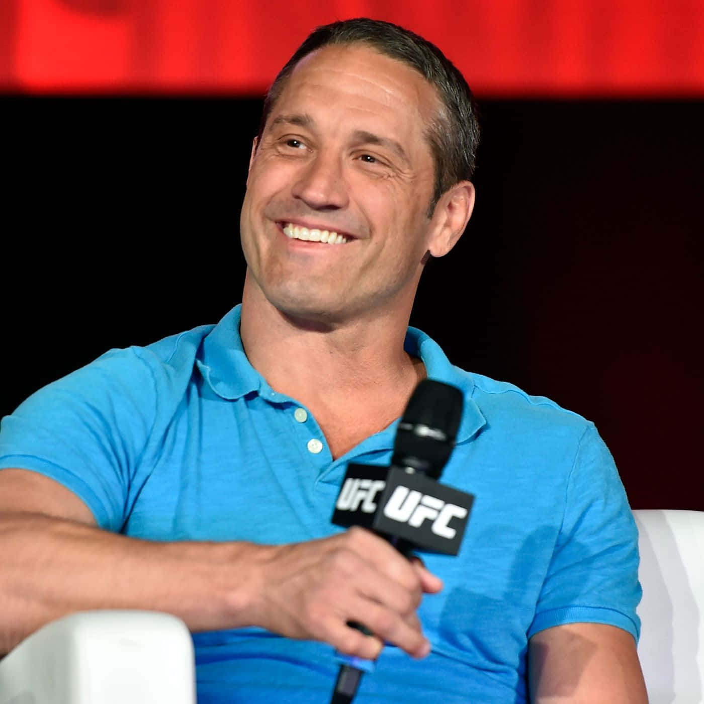UFC選手蓋伊·梅澤格微笑肖像 壁紙 — 一位微笑的男子身穿藍色Polo衫，坐在白色椅子上，手持UFC麥克風，背景為紅色和黑色。
