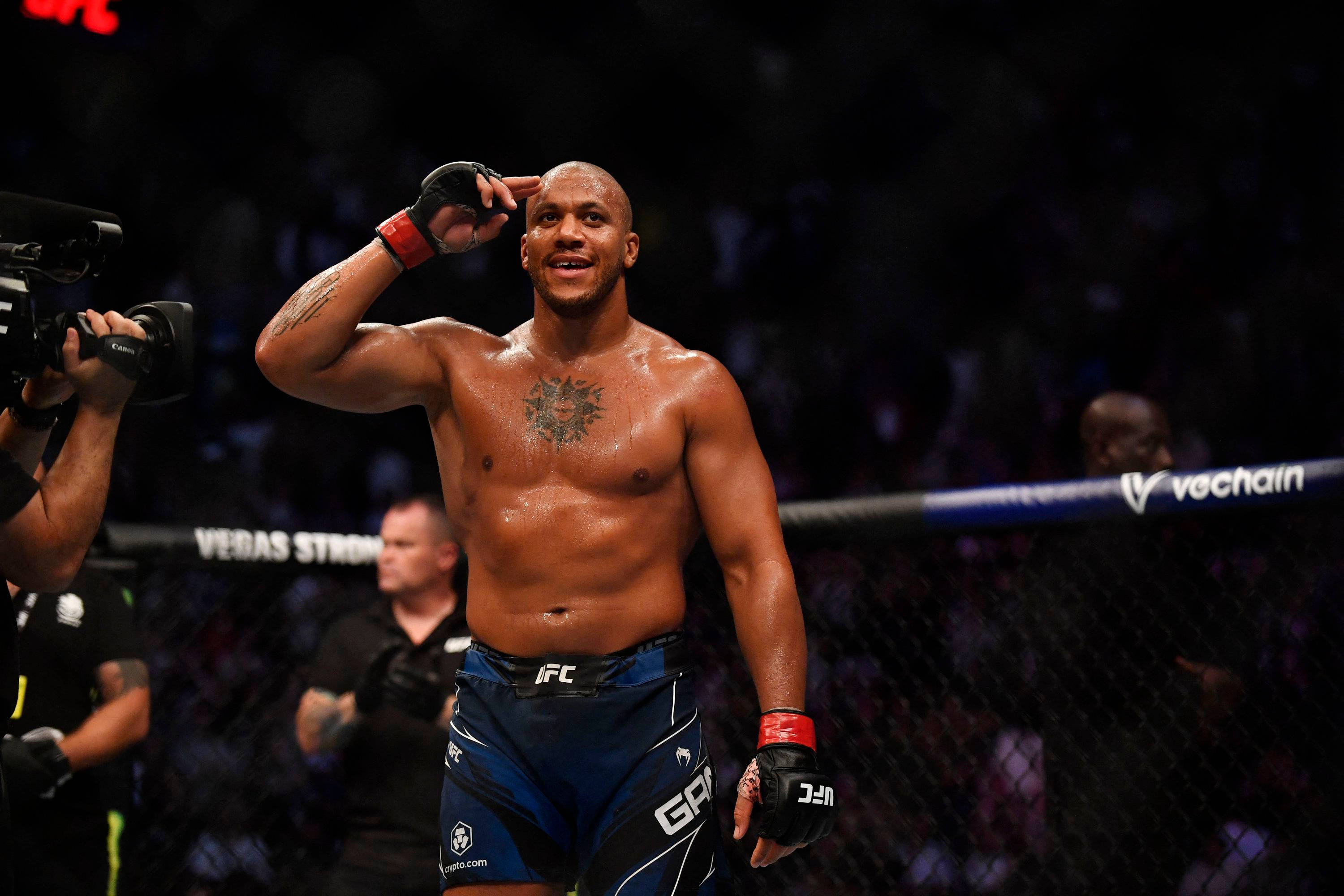 UFC選手西里爾·甘恩在八角籠內敬禮 壁紙 — 一位勝利的混合武術選手在燈光明亮的八角籠內向人群致敬，背景是模糊的觀眾。