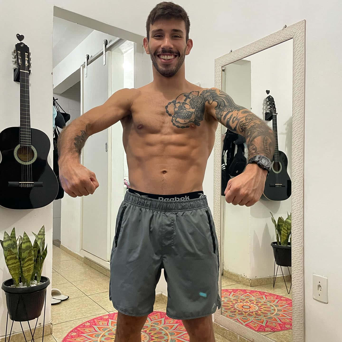 有紋身的肌肉男赤裸上身在一間有結他和鏡子的房間裡展示肌肉。