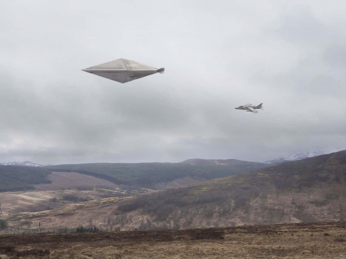 夜空中飛行的神秘UFO 壁紙 — 一艘未來風格的飛碟和一架戰鬥機在陰天籠罩下，飛越崎嶇的山地地形。
