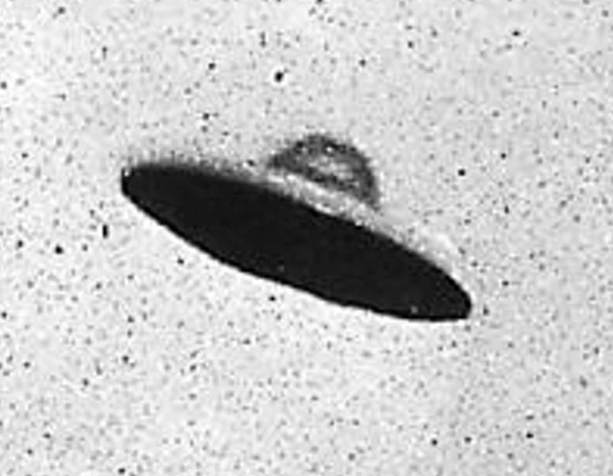 真實 UFO 黑白顆粒感照片 壁紙 — 一張顆粒狀的黑白照片顯示一個黑暗的飛碟在佈滿斑點的天空中盤旋。
