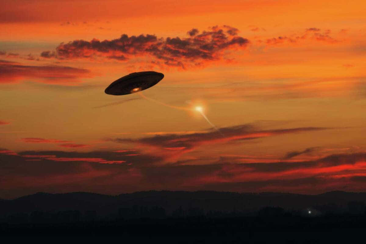 真實UFO飛行於橙色夕陽天空圖 壁紙 — 一個發光的UFO在日落時分懸浮在山脈之上，向天空投射出橙色的光芒。