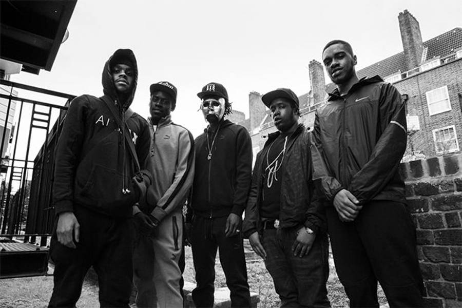 UK Drill 67 男子團體 壁紙 — 五個穿著連帽衫和帽子的年輕男子自信地站在屋頂上，背景是單色磚建築。