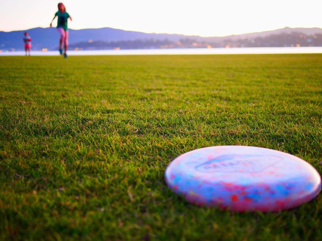 Ultimate Frisbee 中的精彩接盤瞬間 壁紙 — 一個色彩繽紛的飛盤放在鮮綠的草地上，遠處有兩個人在日落時分的湖邊玩耍。